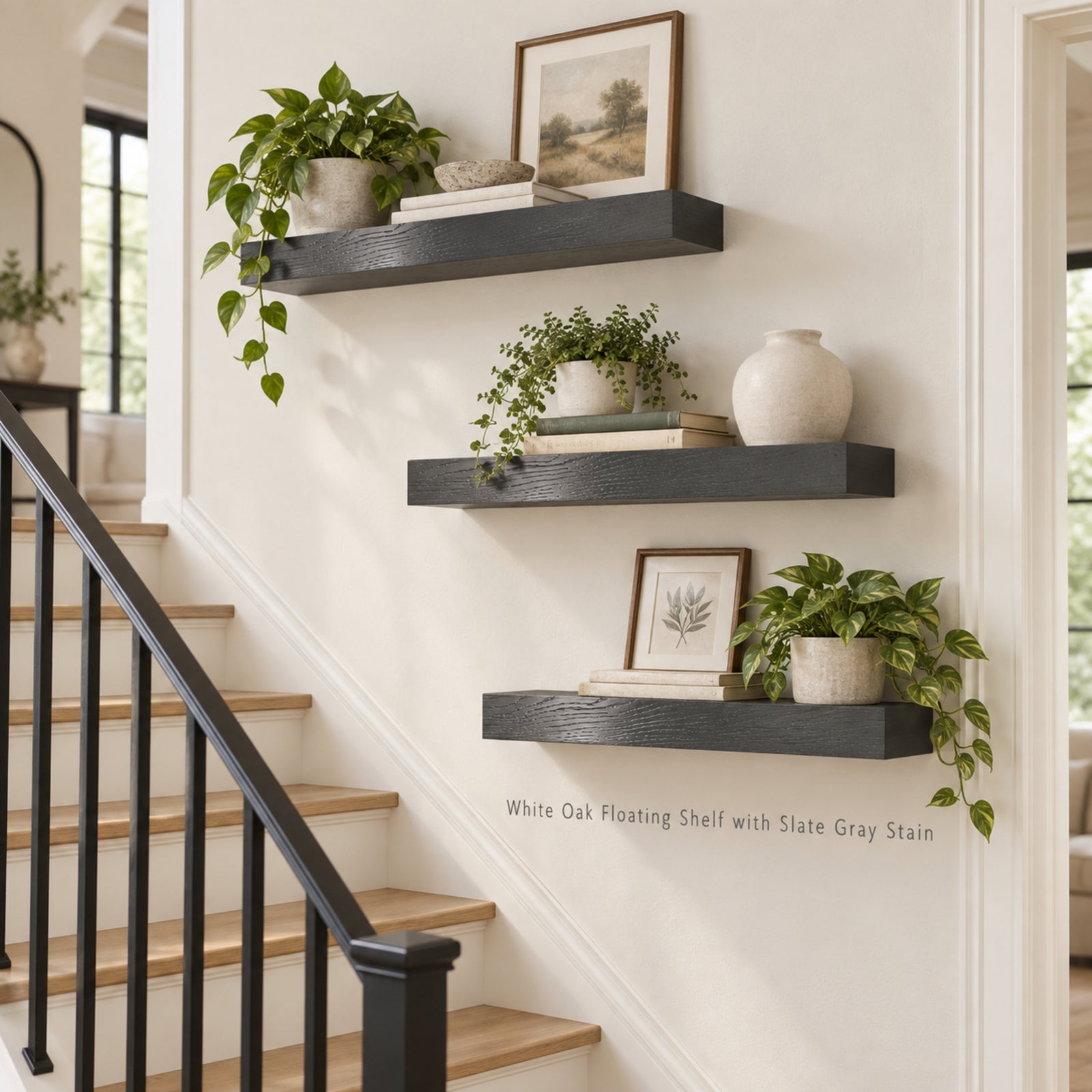 Three floating wooden shelves mounted on a white wall beside a staircase, styled with potted green plants, books, framed art, and a ceramic vase. Text on the wall reads 'White Oak Floating Shelf with Slate Gray Stain'.