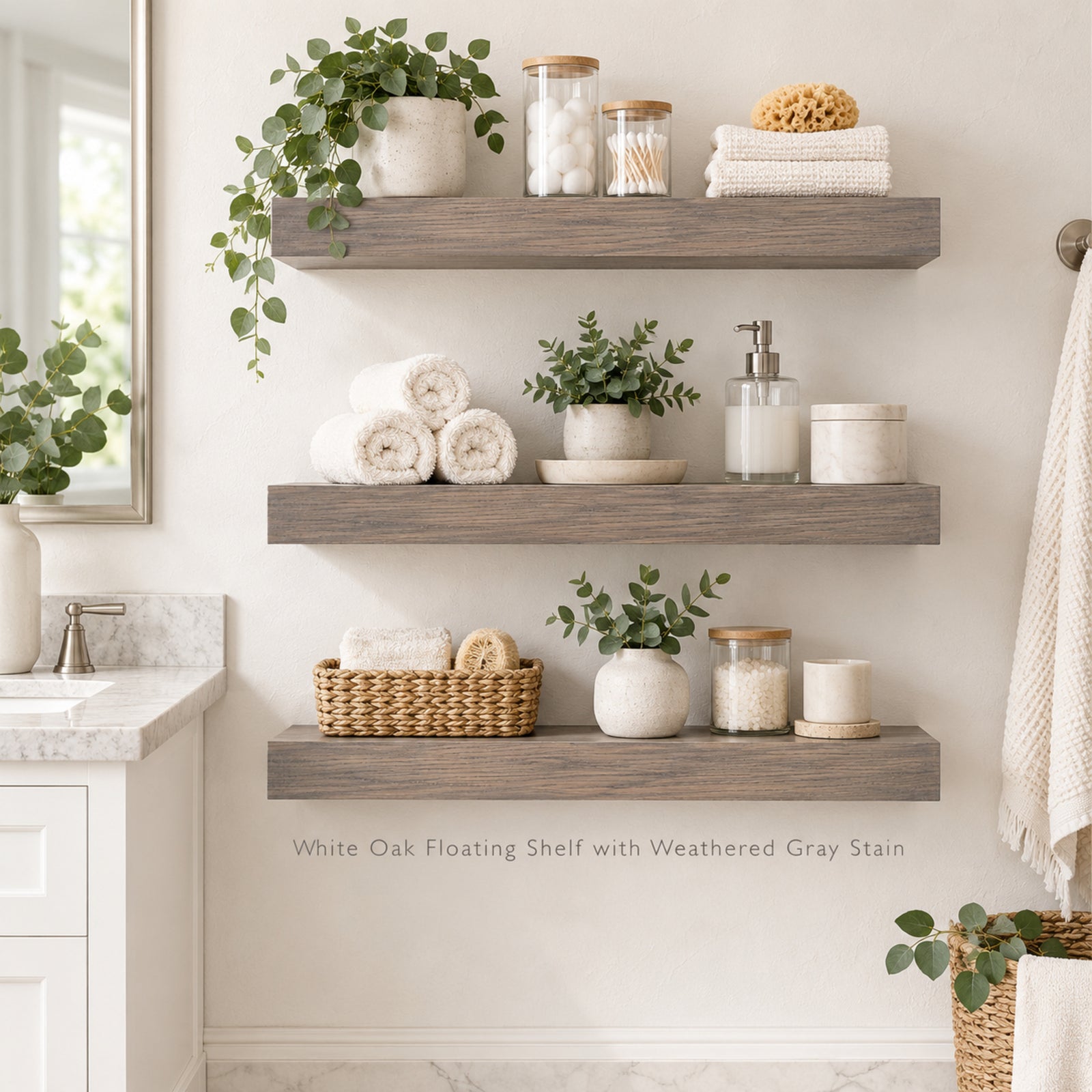 Three floating wall shelves in a bathroom setting, styled with towels, plants, jars, and a basket. Text at the bottom reads 'White Oak Floating Shelf with Weathered Gray Stain'.