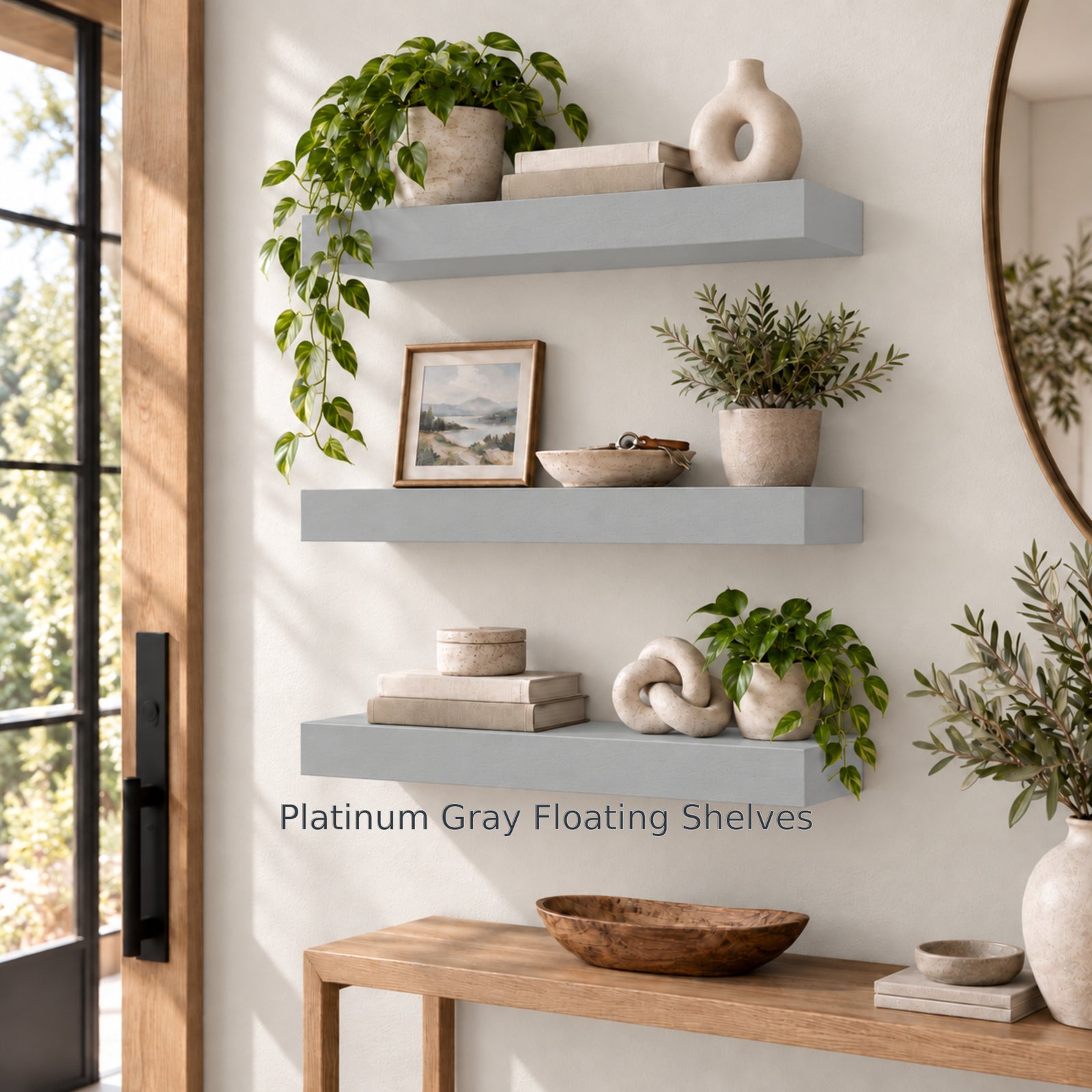 A bright entryway setup featuring three thick platinum gray floating shelves on a light beige wall next to a glass front door. The shelves are decorated with trailing plants, ceramic vases, stacks of books, a small framed landscape print, and a knot sculpture above a wooden console table. Text near the bottom shelf reads "Platinum Gray Floating Shelves".