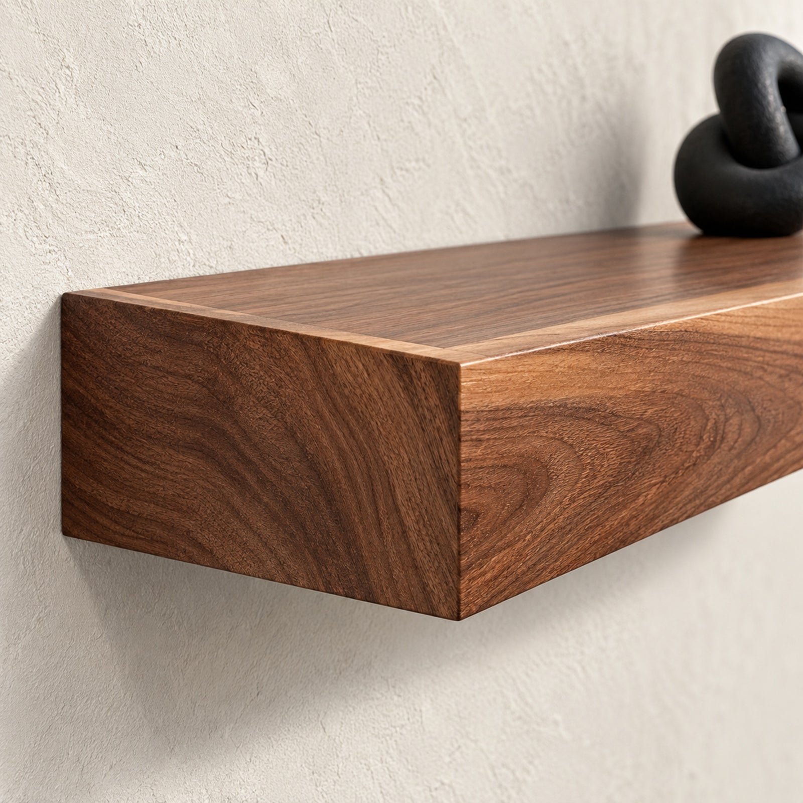Detail image of a floating wooden shelf with a rich walnut grain finish mounted on a textured off-white plaster wall, with a small black decorative object partially visible on top.