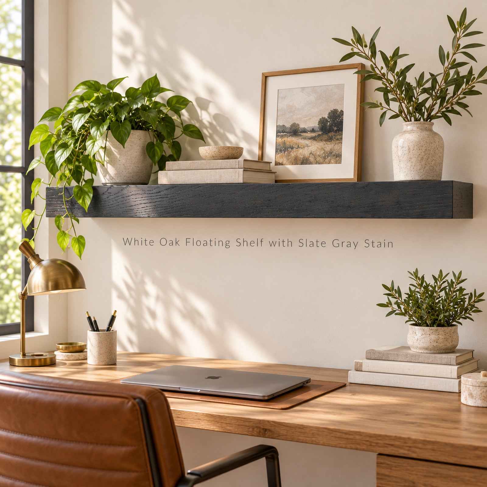 A floating wooden shelf mounted on a white wall above a desk, styled with potted plants, books, a framed print, and a brass desk lamp. The shelf is dark gray stained oak, and the scene is lit by natural sunlight with soft shadows.