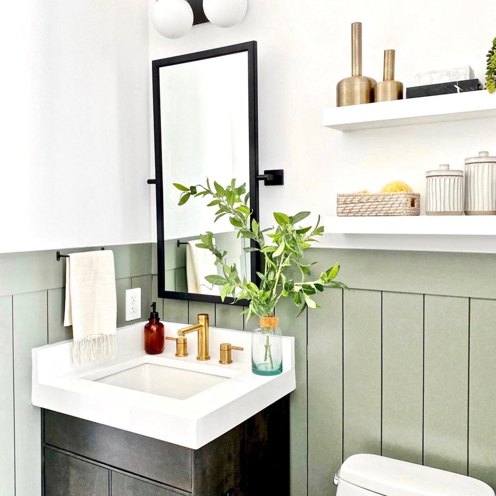 A modern bathroom vanity setup with a white rectangular sink, matte black faucet, and brass fixtures on a dark wood cabinet. A tall black-framed mirror hangs above, with white wall shelves to the right holding decorative vases and jars.