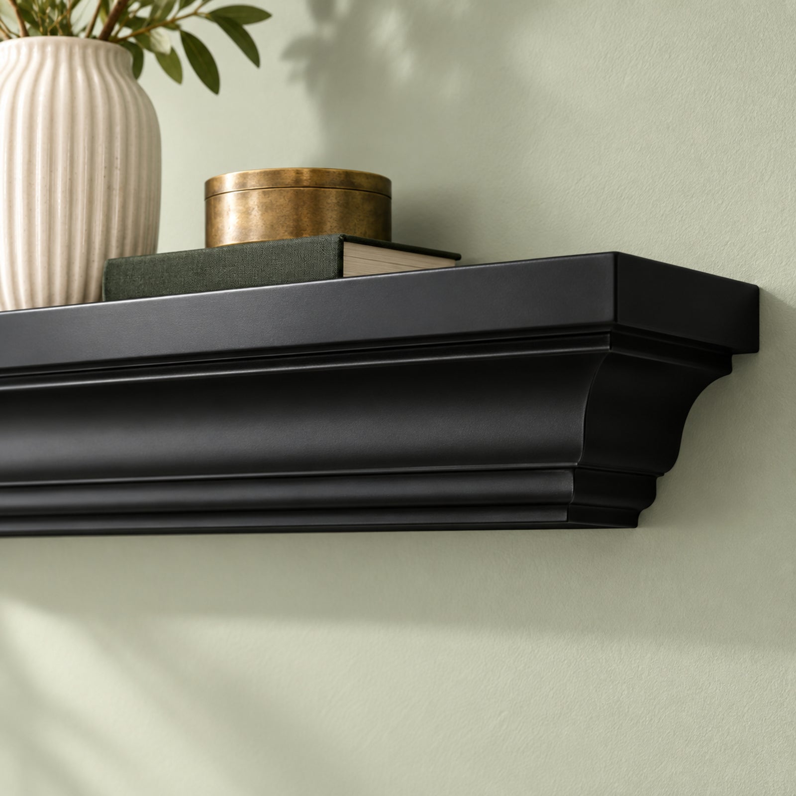 A black painted mantel wall shelf mounted on a light sage green wall, styled with a white ribbed ceramic vase containing green leaves, a dark green book, and a round brass box.