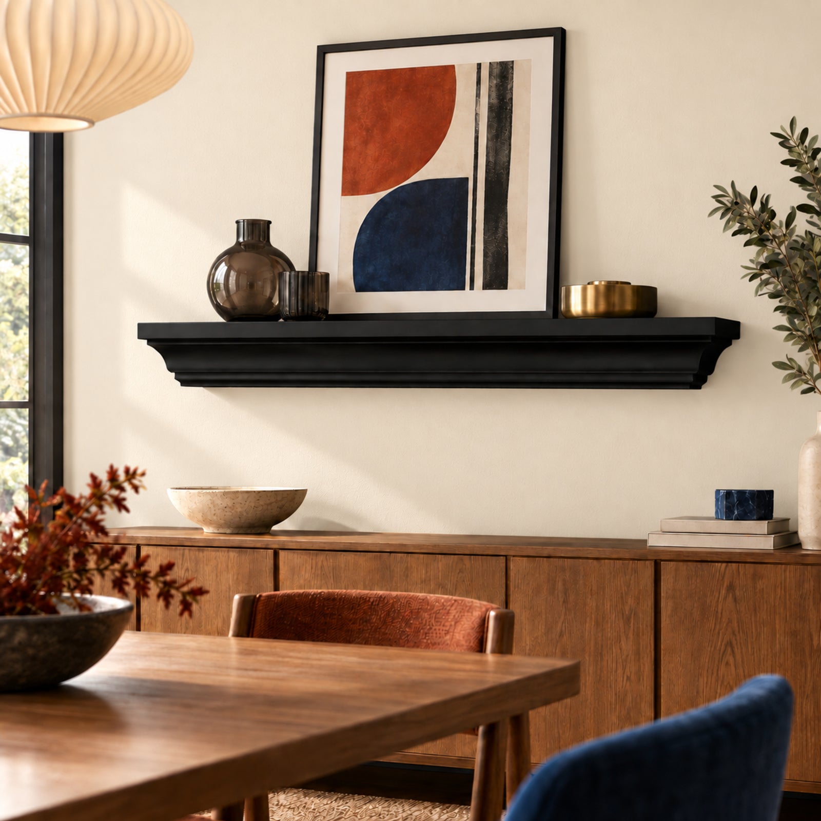 A black floating crown mantel shelf mounted on a beige wall above a wooden credenza, styled with abstract geometric wall art, a dark glass vase, and small gold bowls. In the foreground, a wooden dining table and chairs are partially visible.