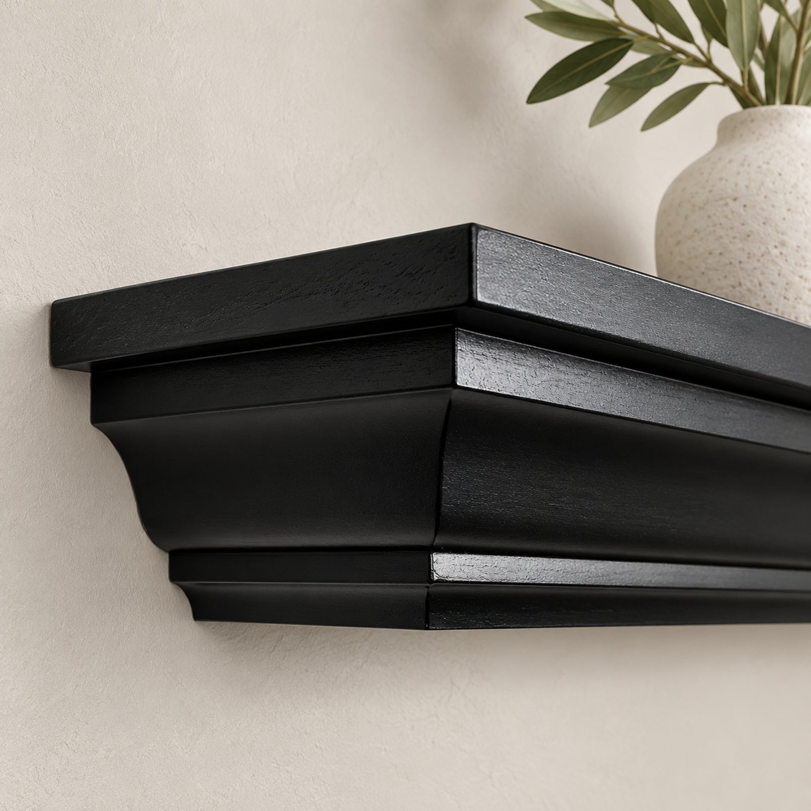 A close-up of a black floating crown mantel shelf mounted on a textured beige wall, with a white ceramic vase and green leaves partially visible on top.