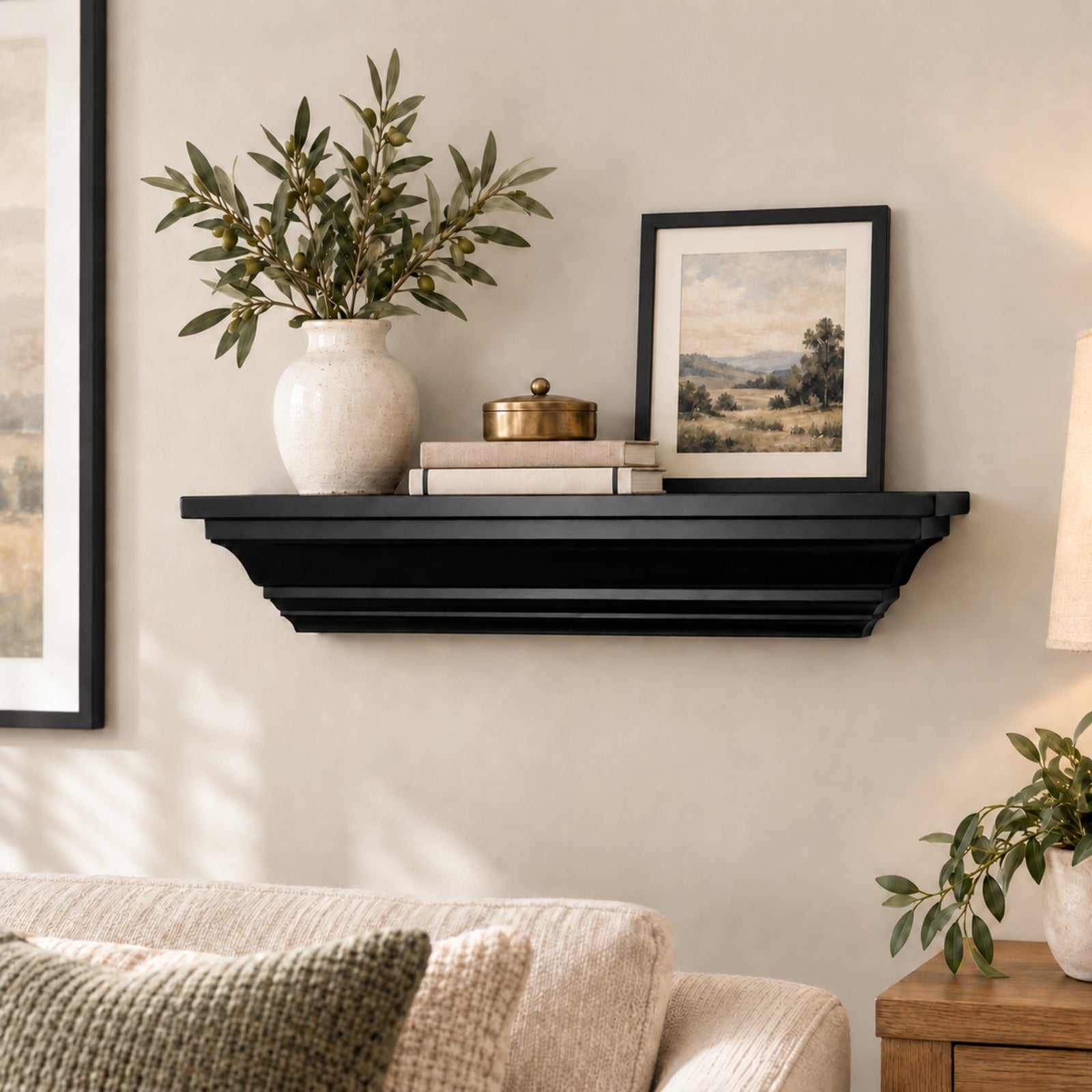 A black floating crown mantel shelf styled in a living room setting, holding a white ceramic vase with green branches, a small brass lidded box, and a framed landscape print. The shelf is mounted on a beige wall above a light beige sofa.