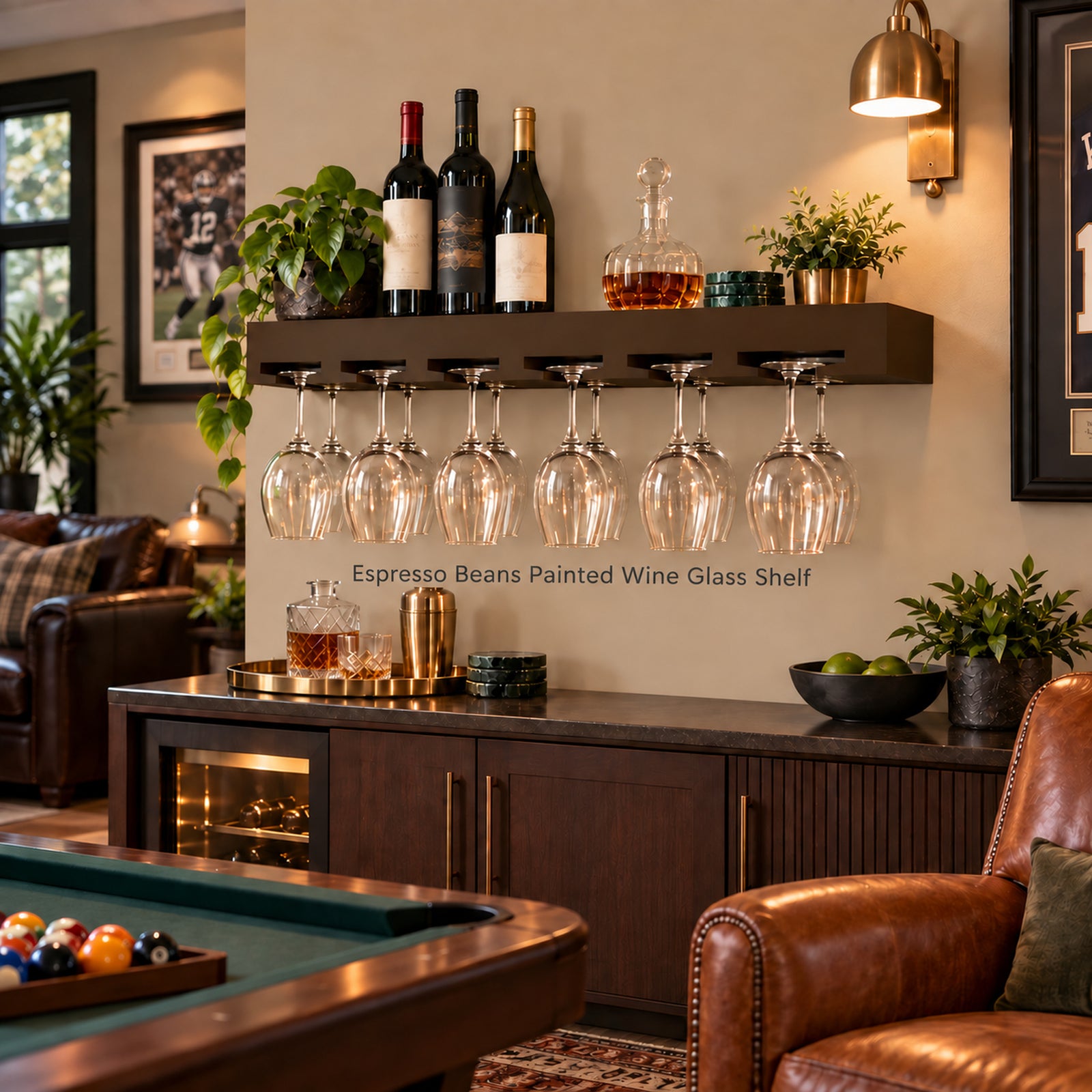 A dark espresso wood wine glass shelf mounted on a beige wall in a home bar setting, holding several wine bottles and multiple wine glasses hanging from a floating rack. The shelf is styled with plants, a decanter, and bar accessories, with a leather chair, pool table, and wall sconce visible in the room.