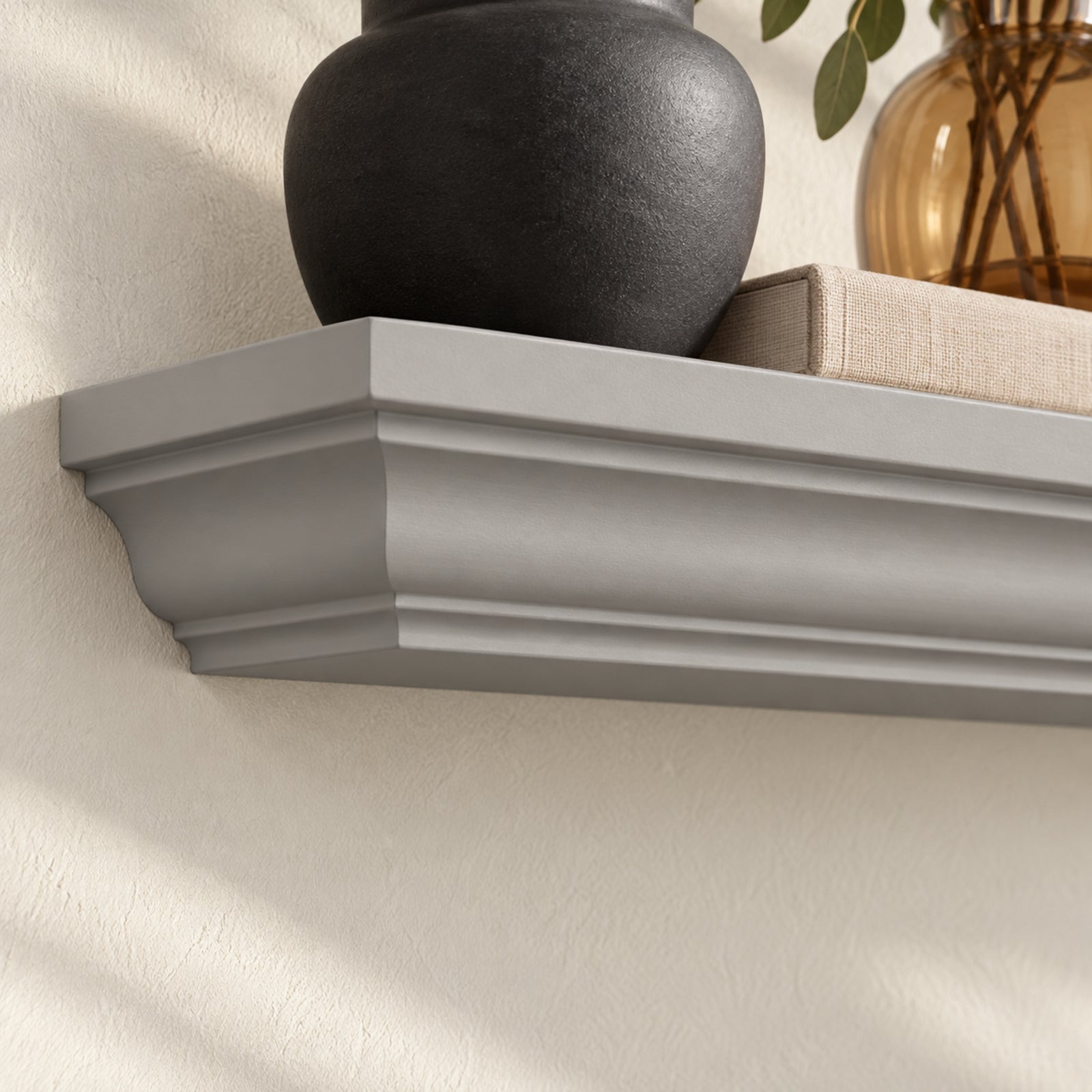 A Harbor gray floating crown mantel shelf mounted on a textured beige wall, styled with a matte black ceramic vase, a beige book, and a glass vase with greenery.