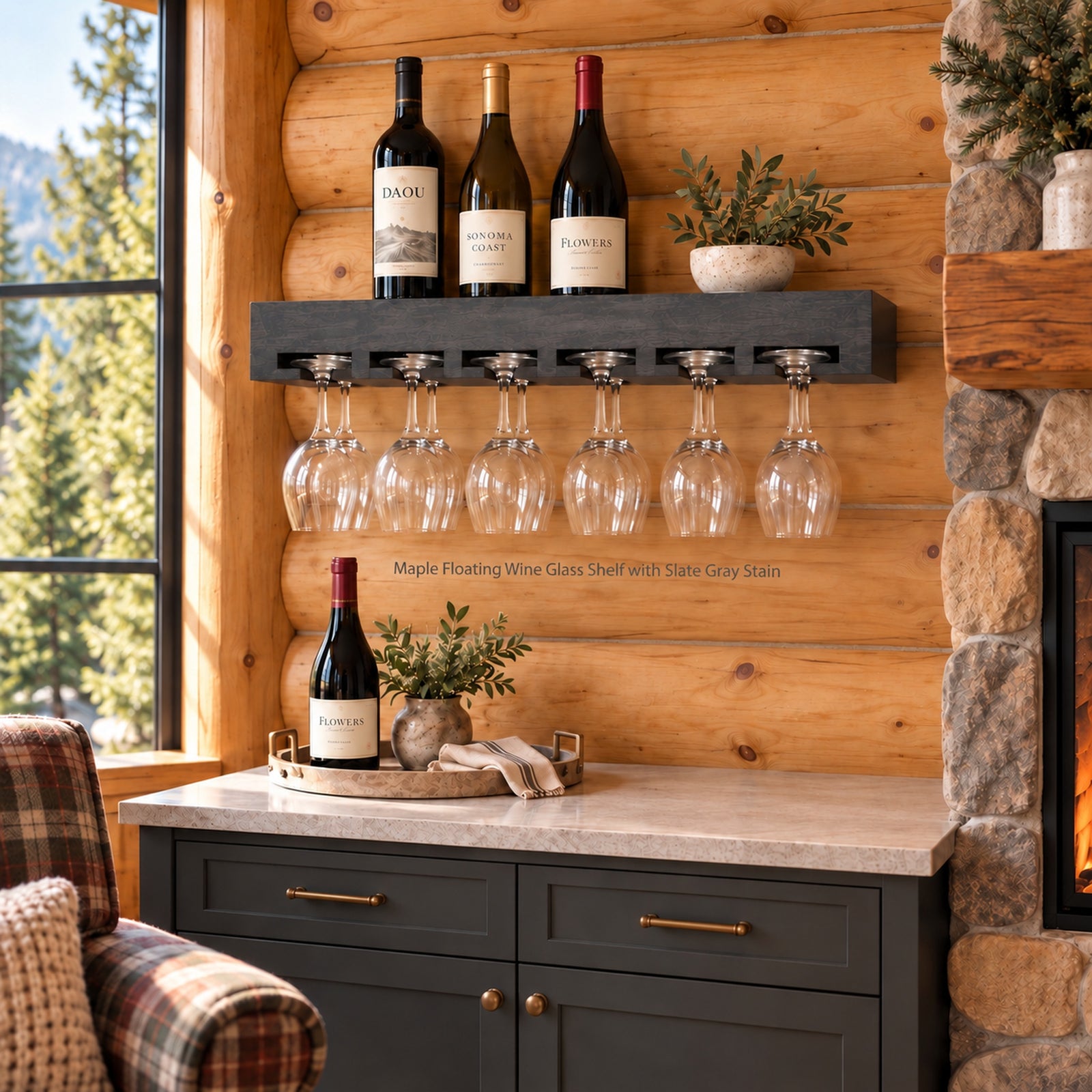 A floating wine glass shelf in a rustic log cabin setting, holding three wine bottles and twelve hanging wine glasses. The shelf is mounted on a wood-paneled wall above a dark gray cabinet with brass handles, next to a stone fireplace and a plaid armchair.