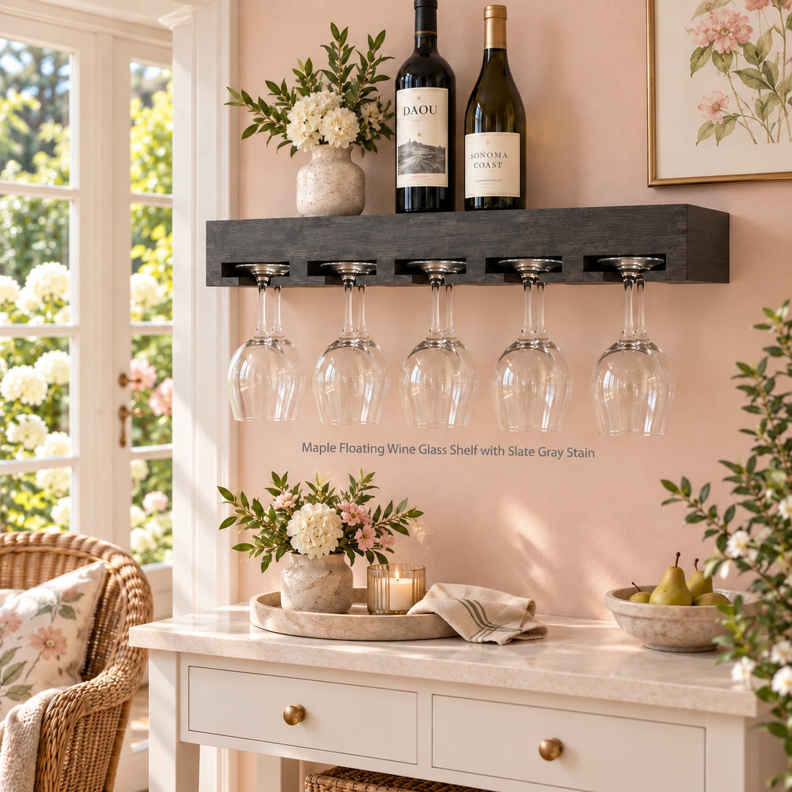 A floating wine glass holder shelf in slate gray stain, mounted on a pink wall above a white console table. The shelf holds two wine bottles and a small vase of flowers, with ten clear wine glasses hanging from the integrated glass holders.