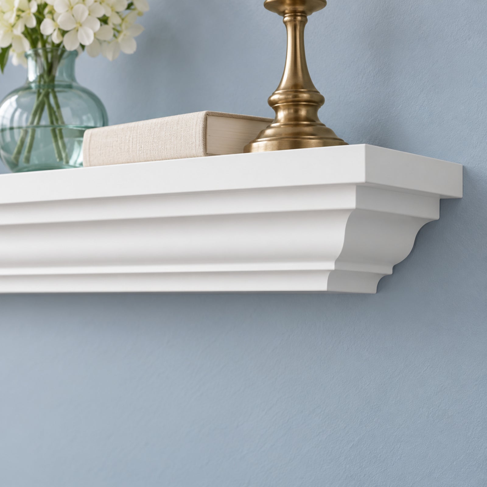 A white floating crown mantel shelf mounted on a light blue wall, styled with a gold candlestick base, a beige book, and a glass vase with white flowers.