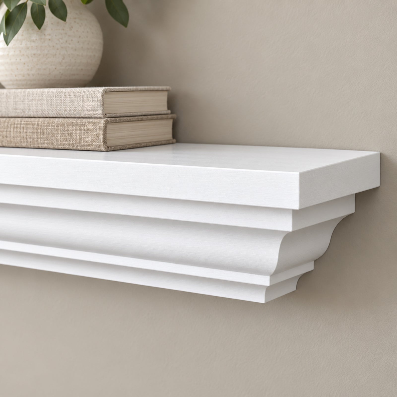 A white floating crown mantel shelf mounted on a beige wall, styled with two stacked books and the top of a textured ceramic vase with green leaves.