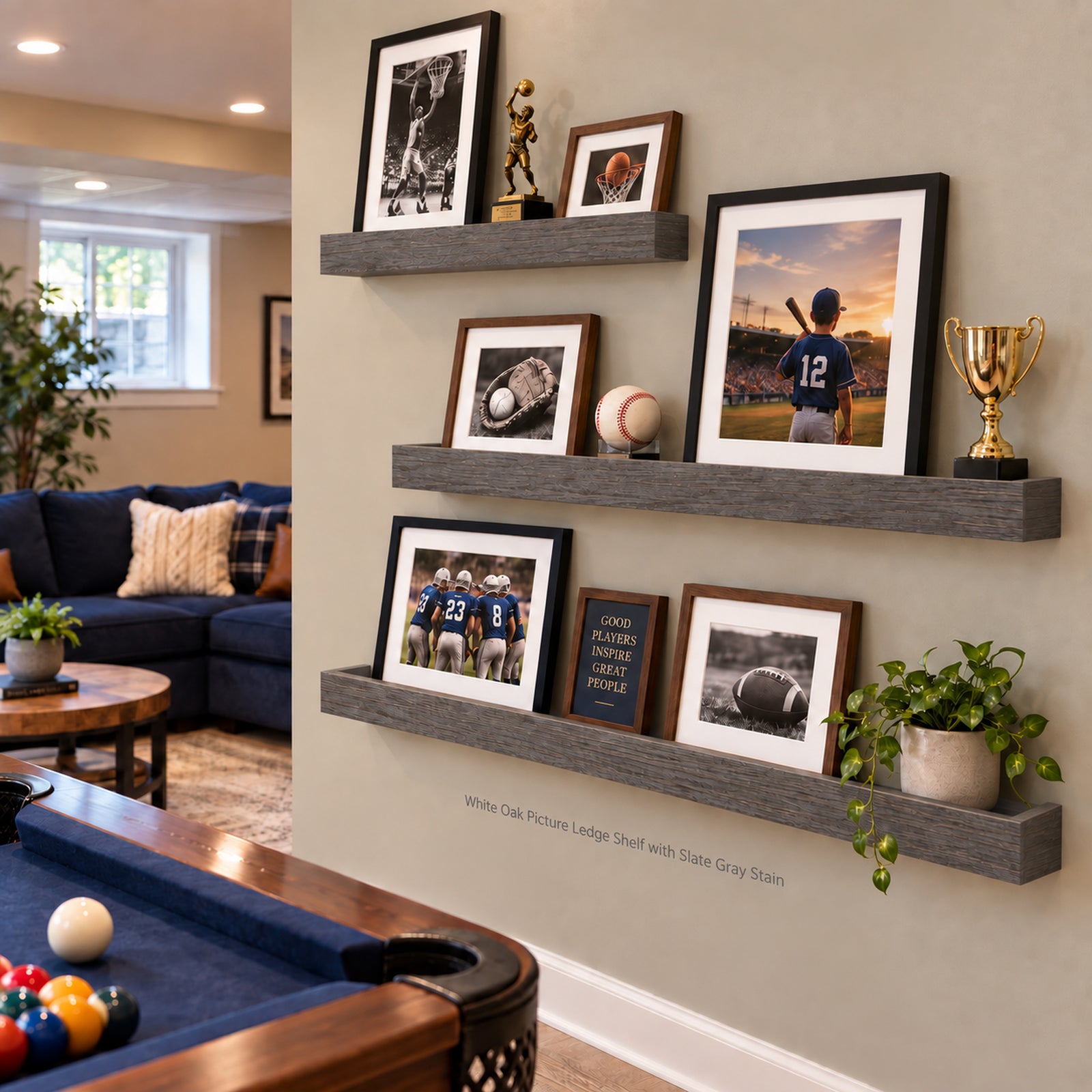 Gray floating ledge shelves mounted on a beige wall in a living room, styled with framed sports photographs, trophies, baseballs, a book, and potted plants. A pool table is visible in the foreground.