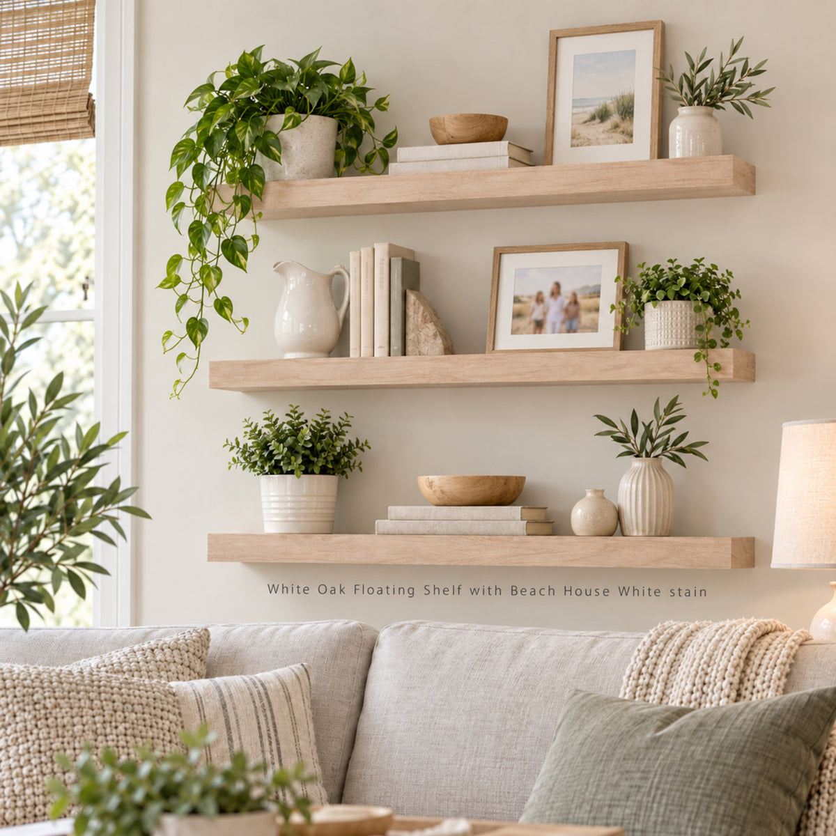 Three white oak floating wall shelves with Beach House White stain styled with books, framed photos, ceramic vases, and potted green plants in a bright living room setting above a beige sofa.