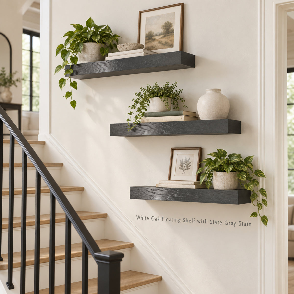 Three floating wooden shelves mounted on a white wall beside a staircase, styled with potted green plants, books, framed art, and a ceramic vase. Text on the wall reads 'White Oak Floating Shelf with Slate Gray Stain'.