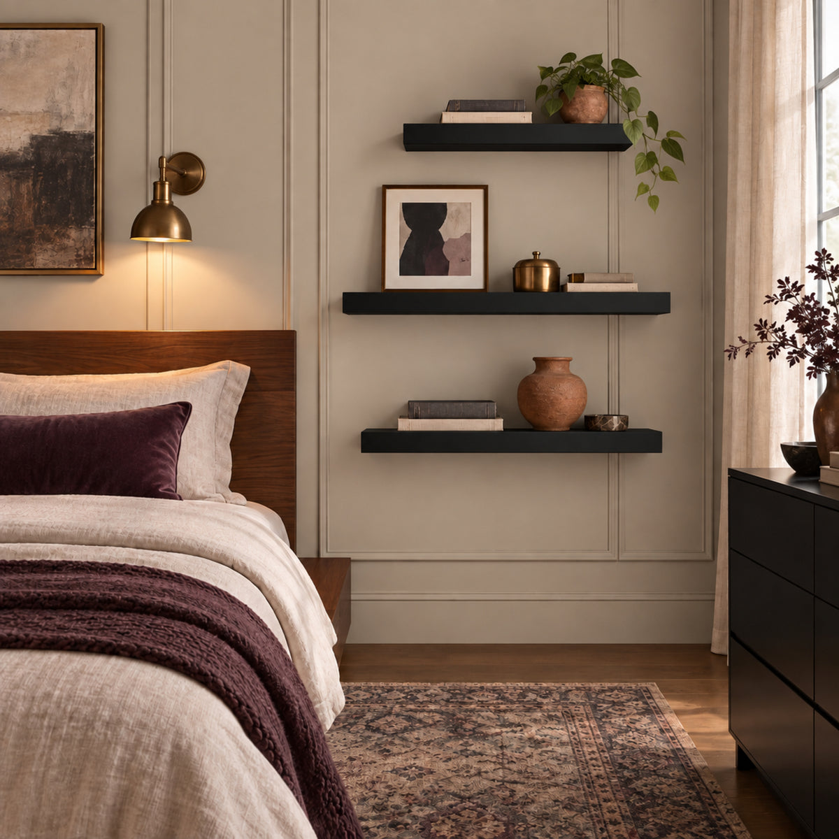 A styled bedroom interior with a bed on the left, three wall-mounted black floating shelves with books, vases, framed art, and a plant, a brass wall lamp, and a patterned rug on a wood floor.