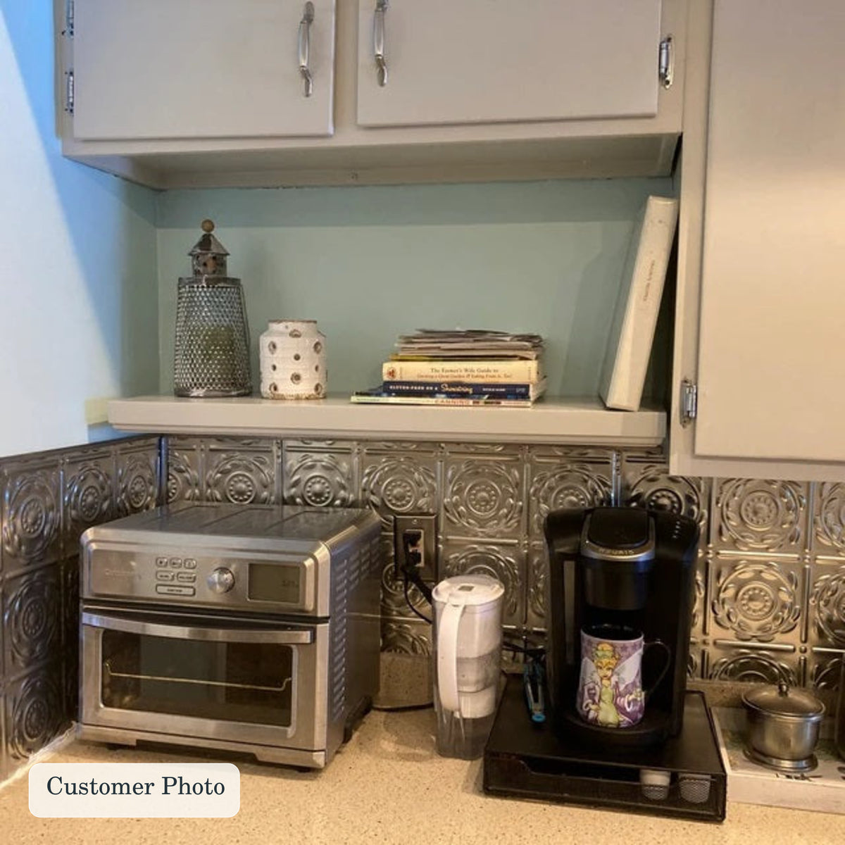 A customer photo of a kitchenette corner showing a stainless steel toaster oven, a black coffee maker with a mug, a water filter pitcher, and a floating shelf with books and a canister against a light blue wall and patterned metal backsplash.