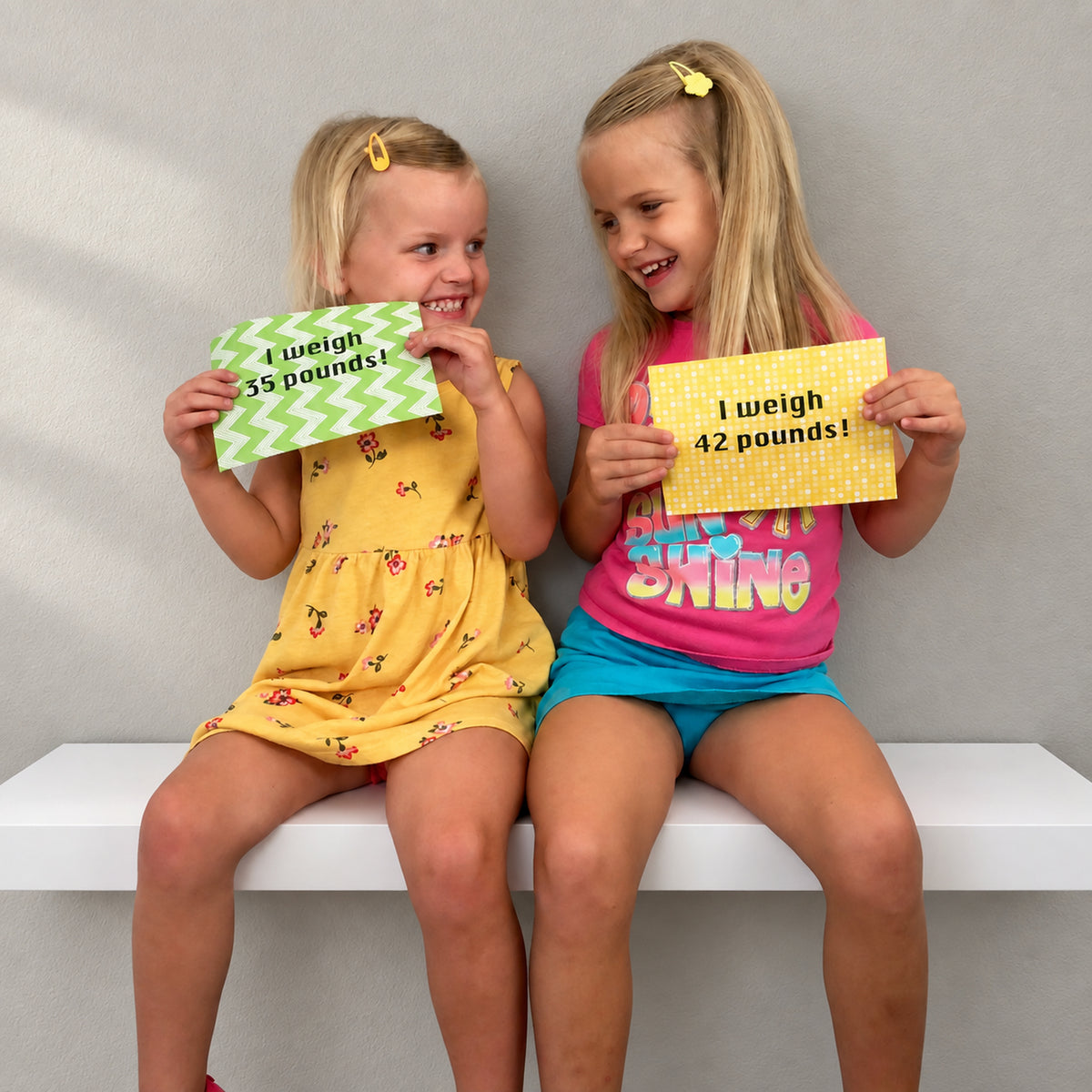 Our granddaughters sitting side-by-side on a deep white floating shelf against a gray wall, holding up paper signs with text that says 'I weigh X pounds!'. The girl on the left wears a yellow floral romper, and the girl on the right wears a pink graphic t-shirt and blue shorts.