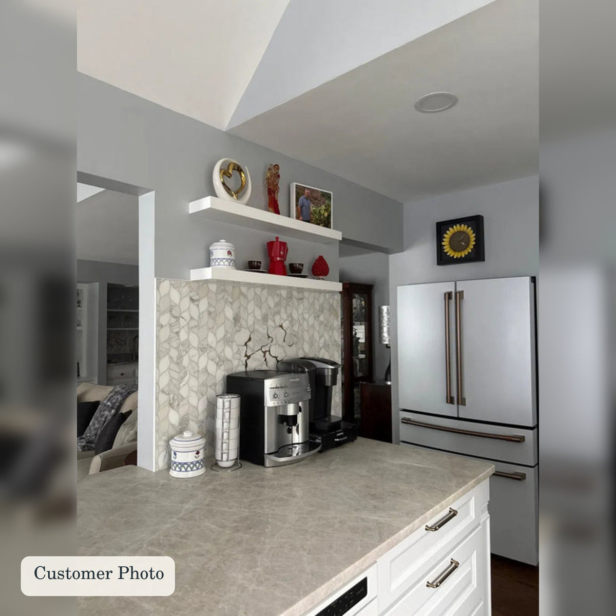 A modern kitchen interior with a stone-look countertop, two white floating shelves with decorative items, a mosaic tile backsplash, a stainless steel refrigerator, and a coffee maker on the counter. 
