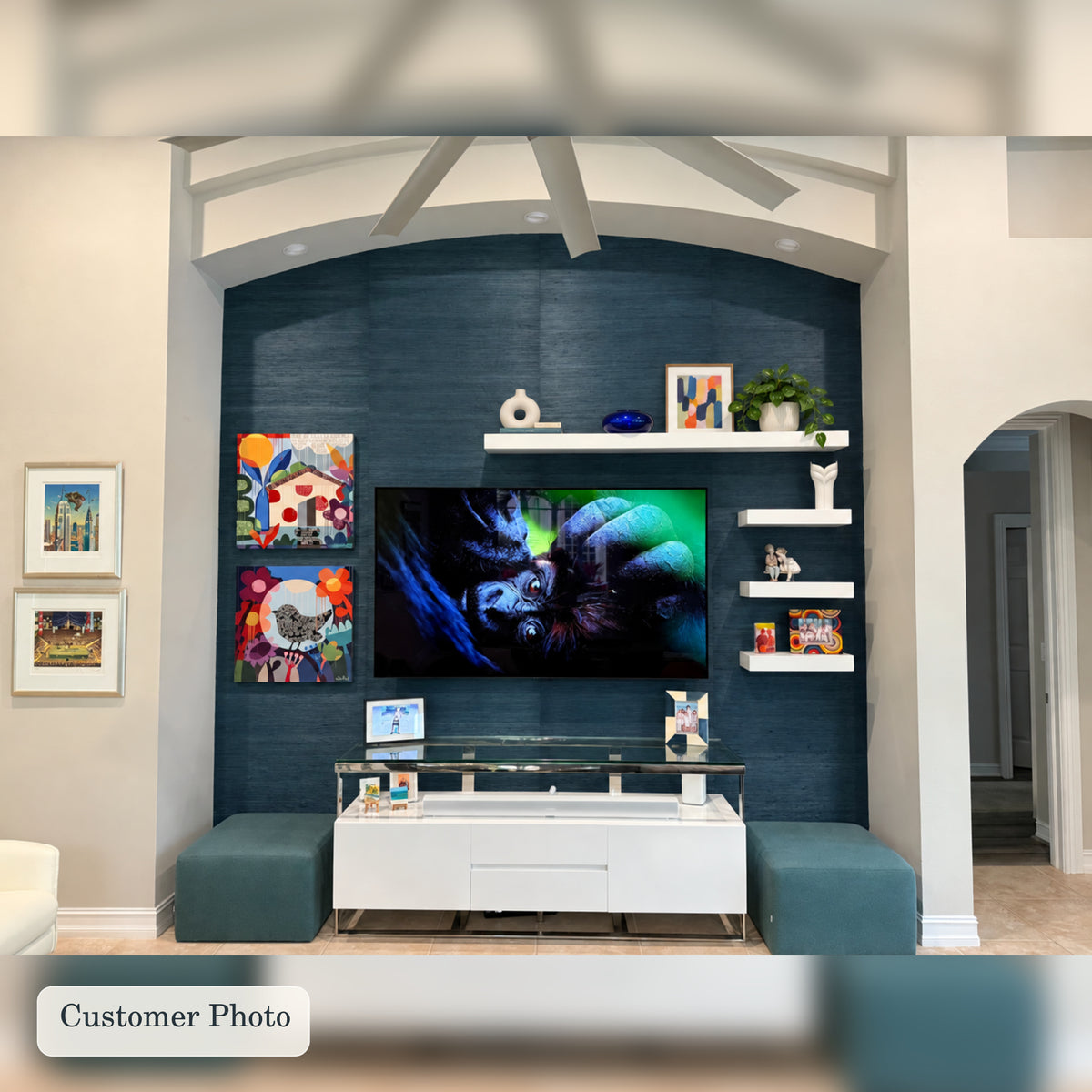 A modern living room setup featuring a dark teal textured accent wall behind a wall-mounted TV, with four white painted floating shelves, colorful framed art, and matching teal ottomans in front of a white media console.