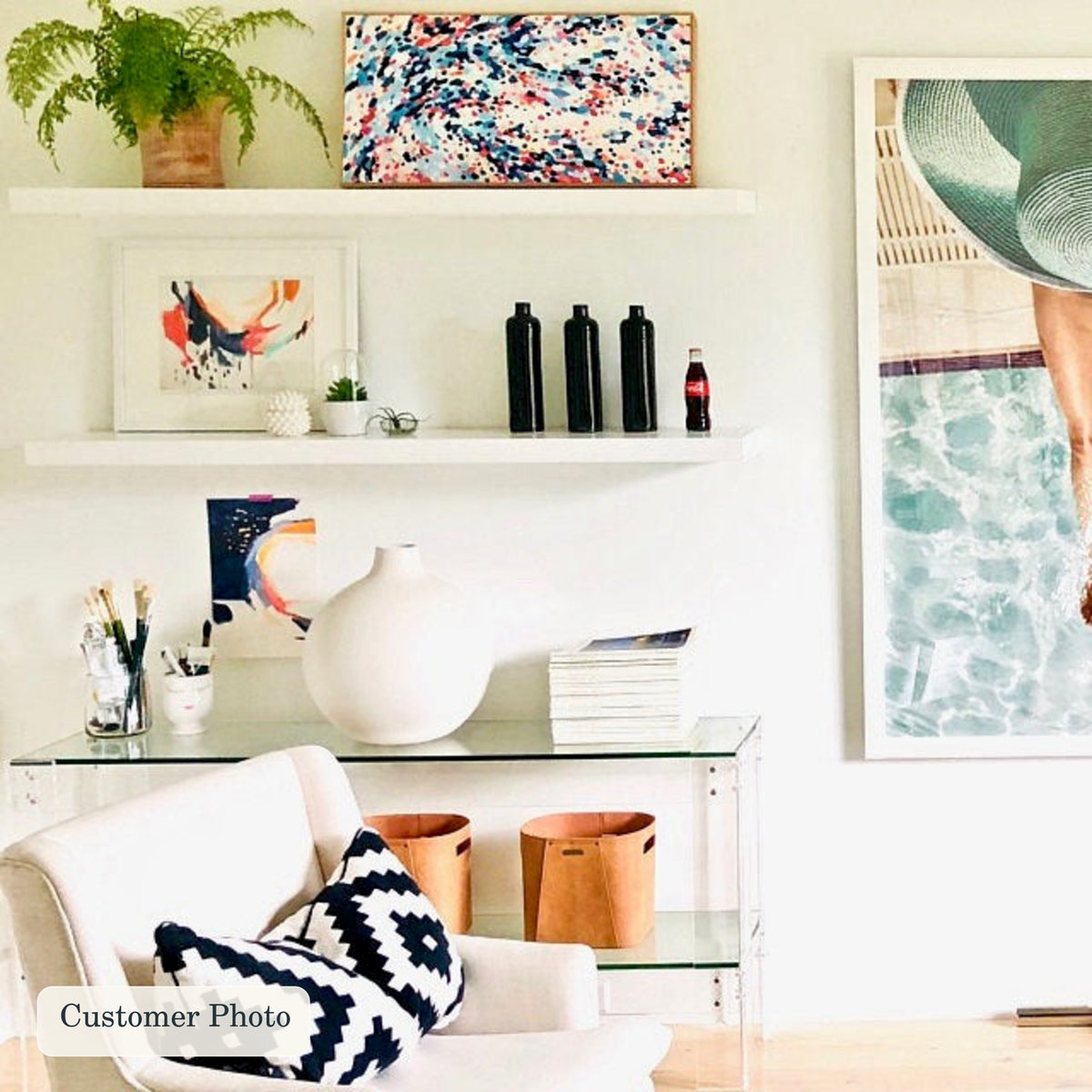 A styled interior scene with two white floating shelves and a glass side table holding decor items, including a white vase, books, and small black bottles. A white armchair with navy-and-white patterned pillows sits in front.