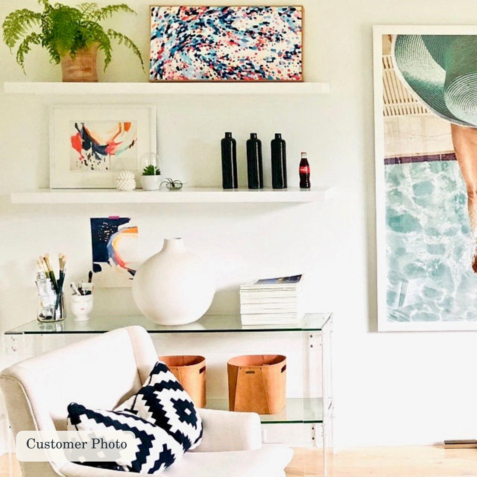 A styled interior scene with two white floating shelves and a glass side table holding decor items, including a white vase, books, and small black bottles. A white armchair with navy-and-white patterned pillows sits in front.