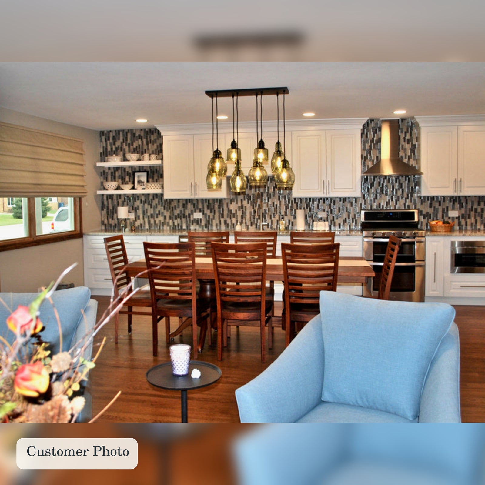 A lifestyle interior photo of a modern kitchen and dining area, featuring a wooden dining table and chairs, a multi-light pendant chandelier, white cabinetry and two white floating shelves, with a mosaic tile backsplash. 
