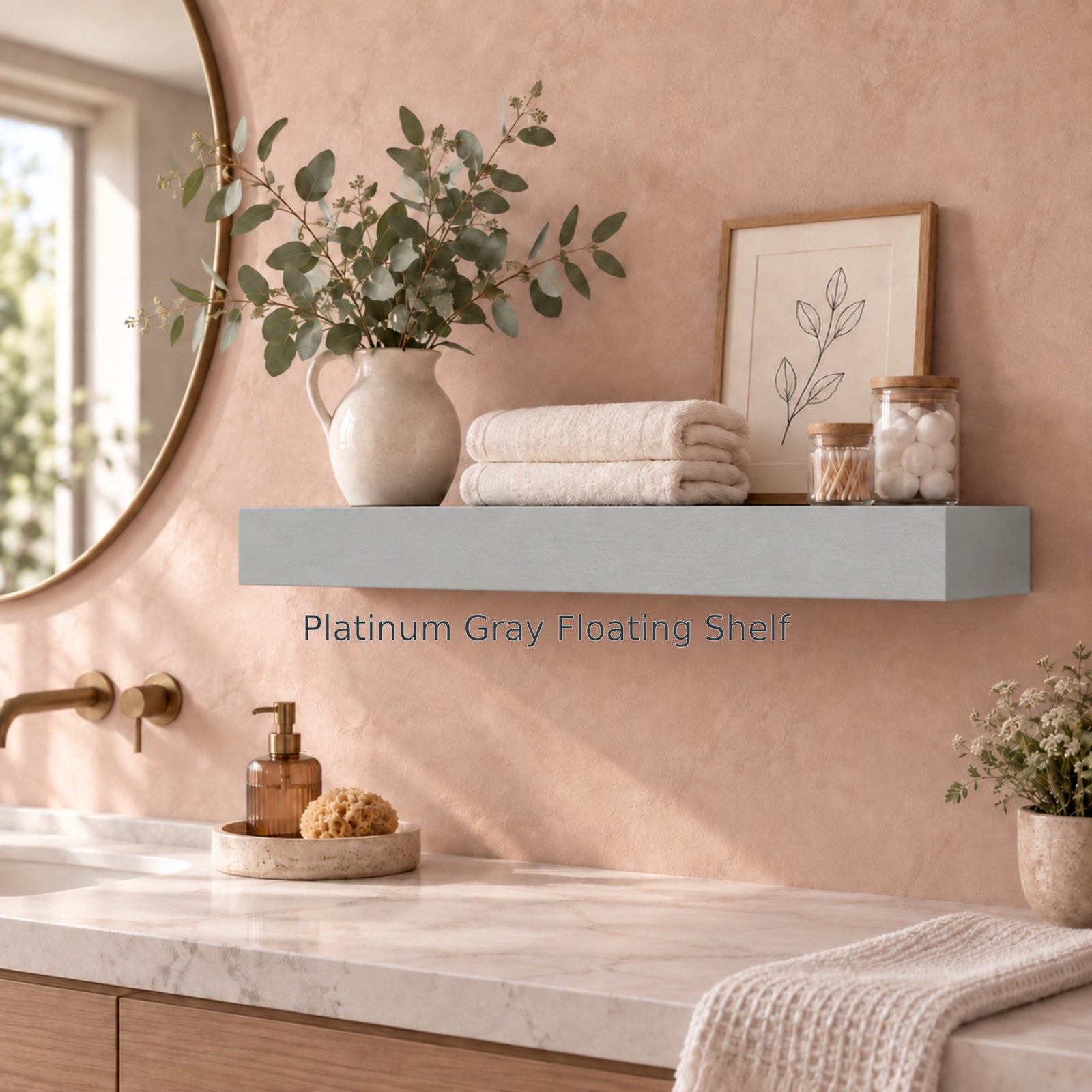 A serene bathroom scene featuring a single thick platinum gray floating shelf against a warm terracotta plaster wall. The shelf holds folded white hand towels, a pitcher filled with eucalyptus branches, a framed botanical line art print, and glass bathroom jars. Text below the shelf reads "Platinum Gray Floating Shelf".