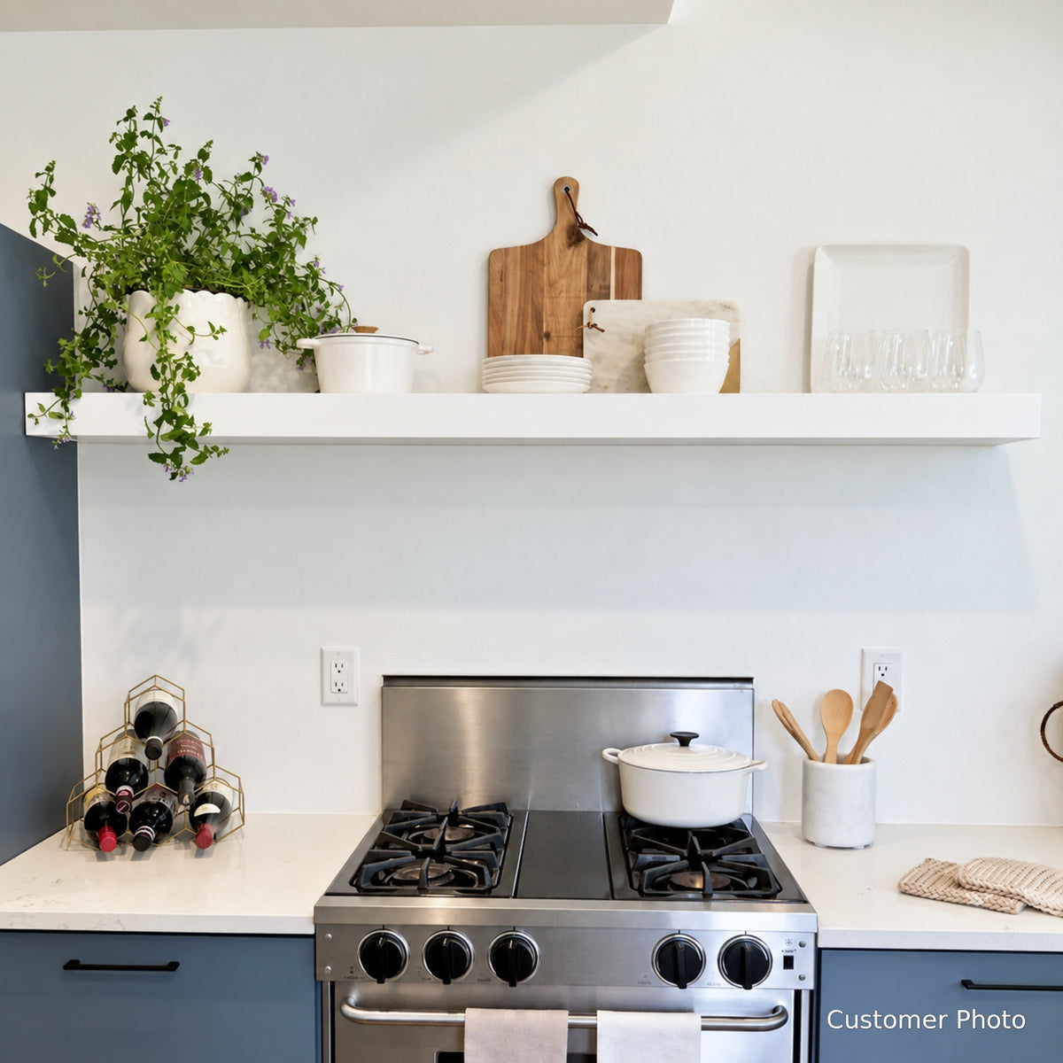 A white floating shelf mounted on a kitchen backsplash above a stove, styled with potted plants, cutting boards, and glassware.