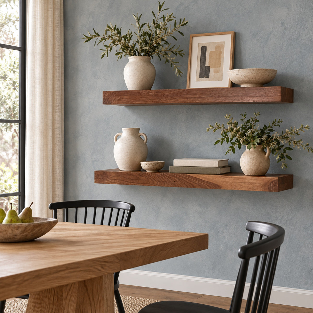 Two floating wooden shelves mounted on a textured blue-gray wall in a dining room setting, styled with ceramic vases, books, a bowl, and framed art.