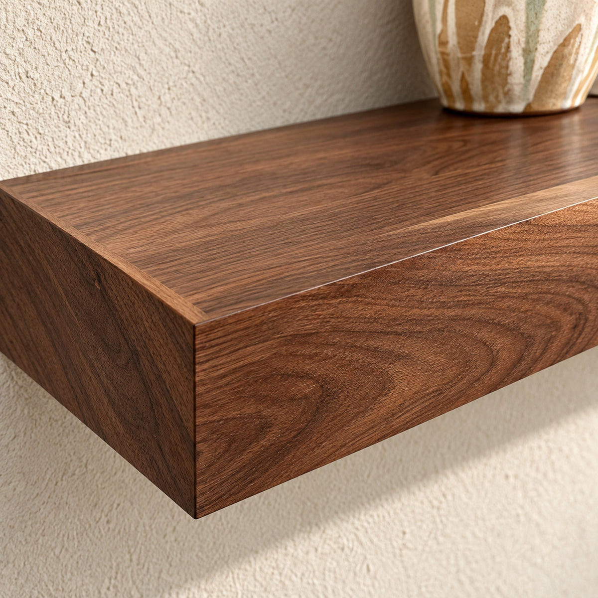 A close-up of a floating wooden shelf with a rich dark brown walnut grain finish, mounted on a textured beige wall with a ceramic vase partially visible at the top right.