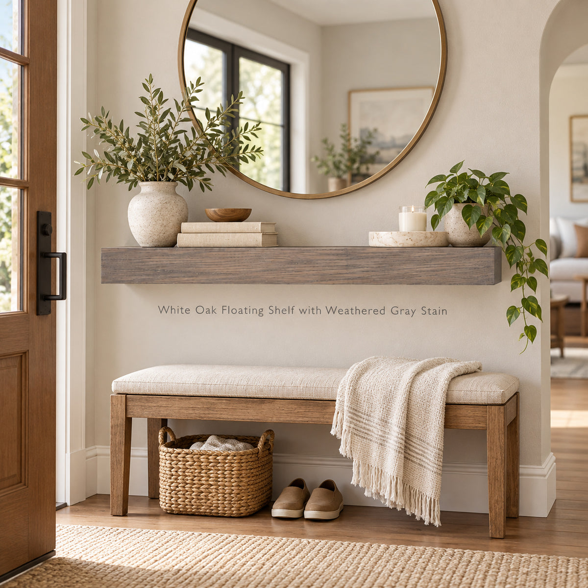A white oak floating shelf with a weathered gray stain is mounted on a beige wall above a wooden bench with a cream upholstered seat in the entryway of a home. The shelf is styled with a ceramic vase of greenery, books, a candle, and a small bowl.