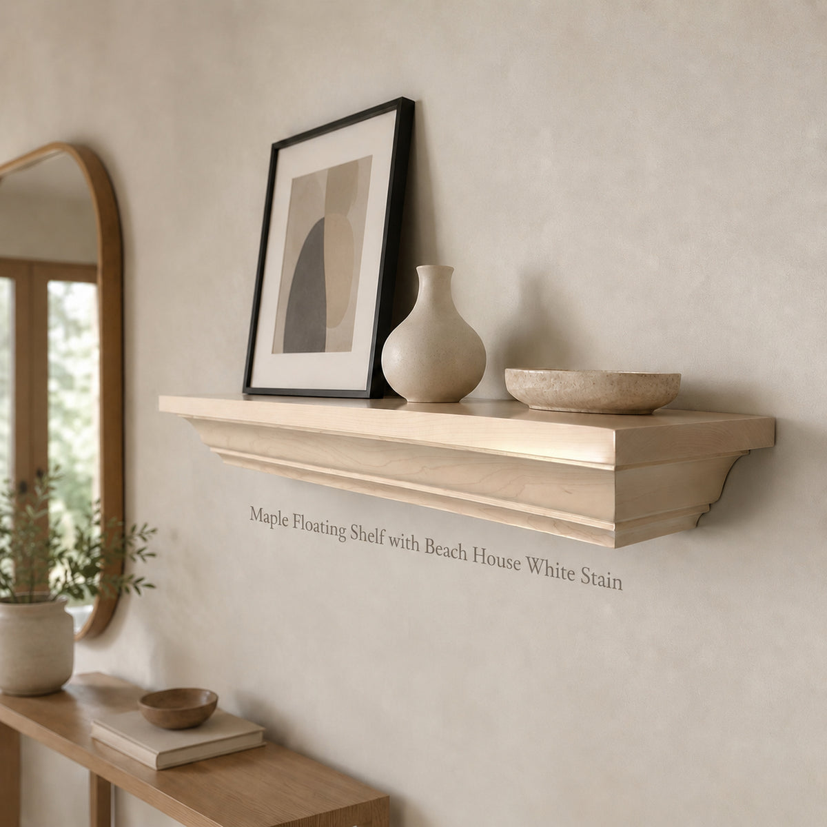 A light natural wood floating shelf mounted on a textured beige wall, styled with a black-framed abstract print, a cream ceramic vase, and a matching wooden bowl. The text 'Maple Floating Shelf with Beach House White Stain' is printed on the wall below the shelf.