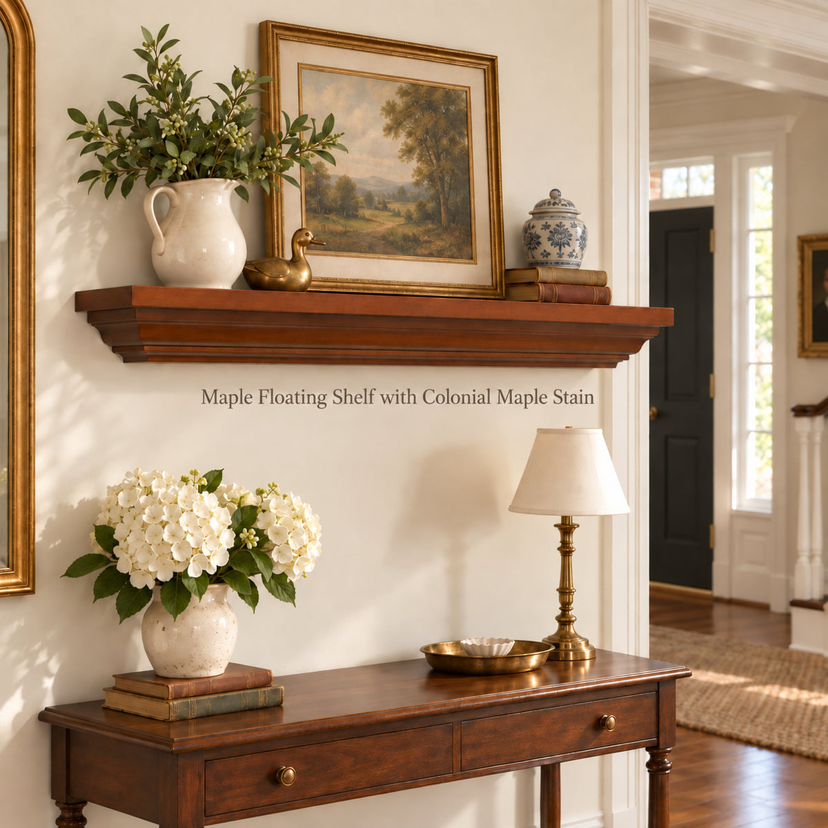 A warm medium-brown floating crown mantel shelf with a Colonial maple finish, styled in a classic entryway above a wooden console table. The shelf is decorated with a white pitcher of greenery, a landscape painting in a gold frame, and a small blue-and-white ceramic jar.