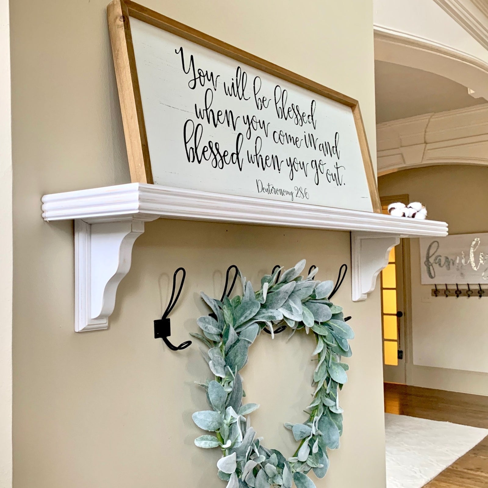 A framed wall sign with a biblical blessing in black script on a white background, displayed on a white farmhouse shelf in a home entryway. The shelf is mounted on a beige wall with black coat hooks below holding a green eucalyptus wreath.