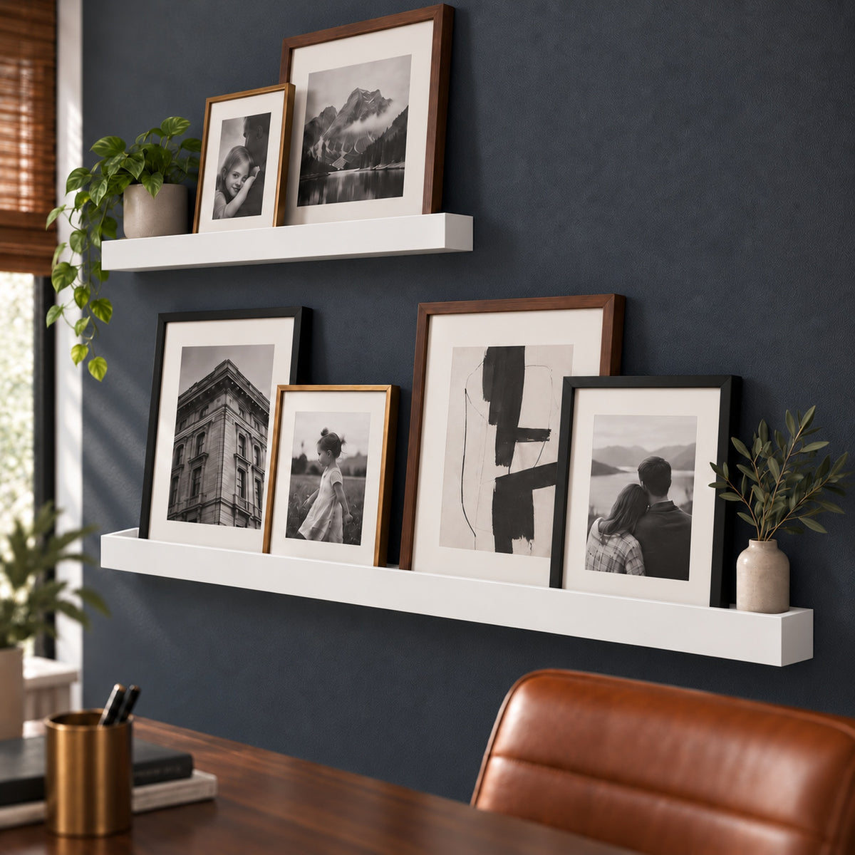 A styled home office scene showing two white floating ledge shelves mounted on a dark blue wall, each holding several framed black-and-white and brown photos. A small potted plant and a vase with greenery sit on the shelves, with part of a wooden desk and a brown leather chair visible in the foreground.