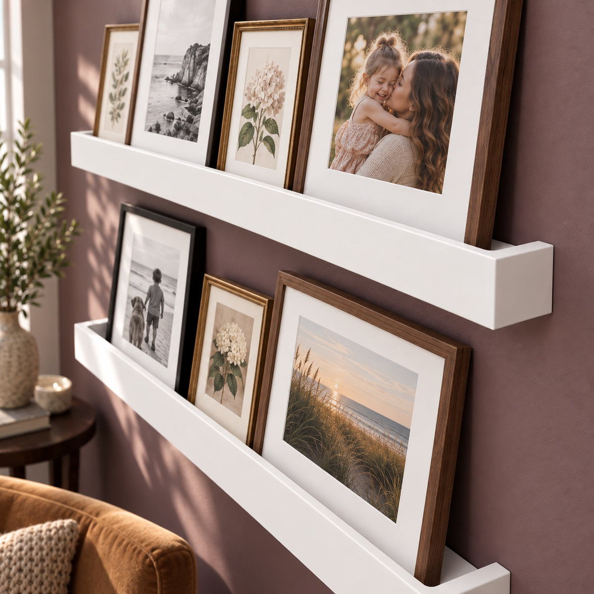 Two white floating ledge shelves mounted on a mauve wall, styled with multiple framed family photographs and botanical prints. A portion of a brown velvet armchair and a potted plant are visible in the foreground and left side.