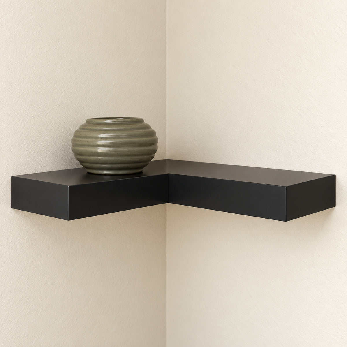 A modern black L-shaped floating corner shelf mounted on two cream-colored textured walls, holding a ribbed olive-green ceramic vase.