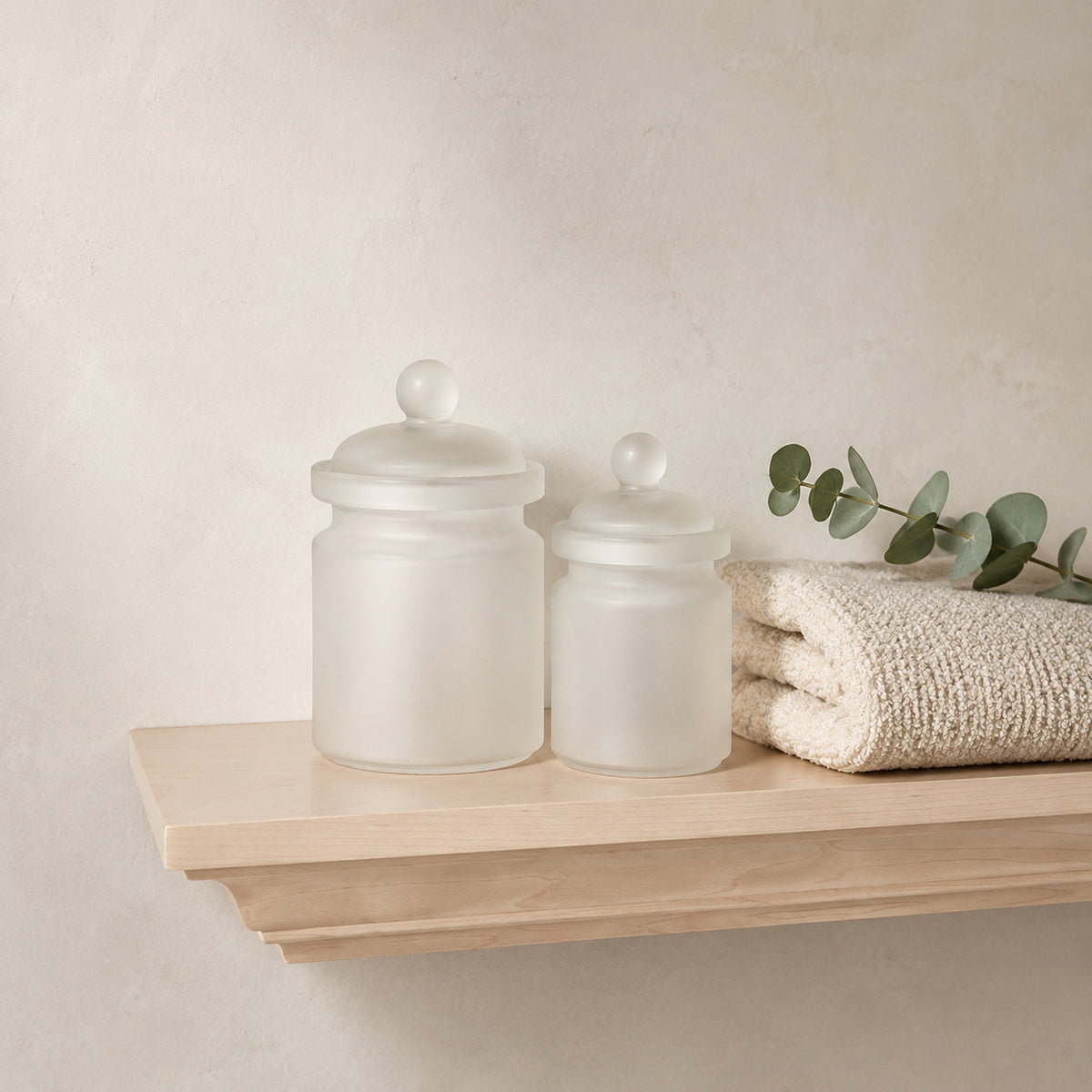 Two frosted glass canisters with matching lids sit on a light wood floating shelf beside a folded cream woven textile and a green eucalyptus sprig, in a modern bathroom.