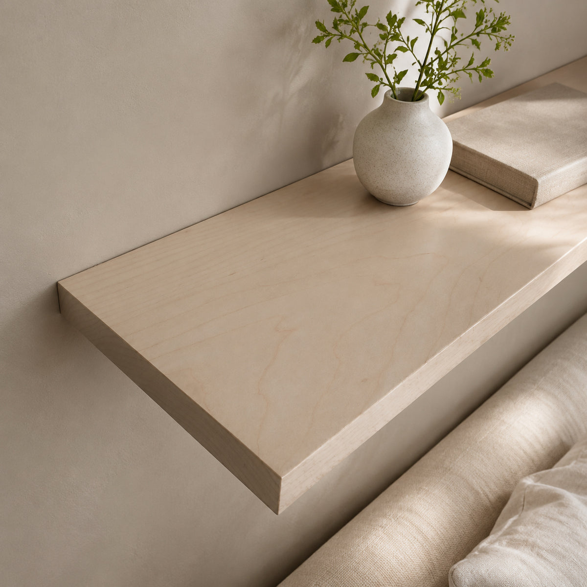 A light beige floating shelf mounted on a neutral wall, styled with a small textured white vase holding green stems and a closed book, with part of a beige upholstered chair visible in the foreground.