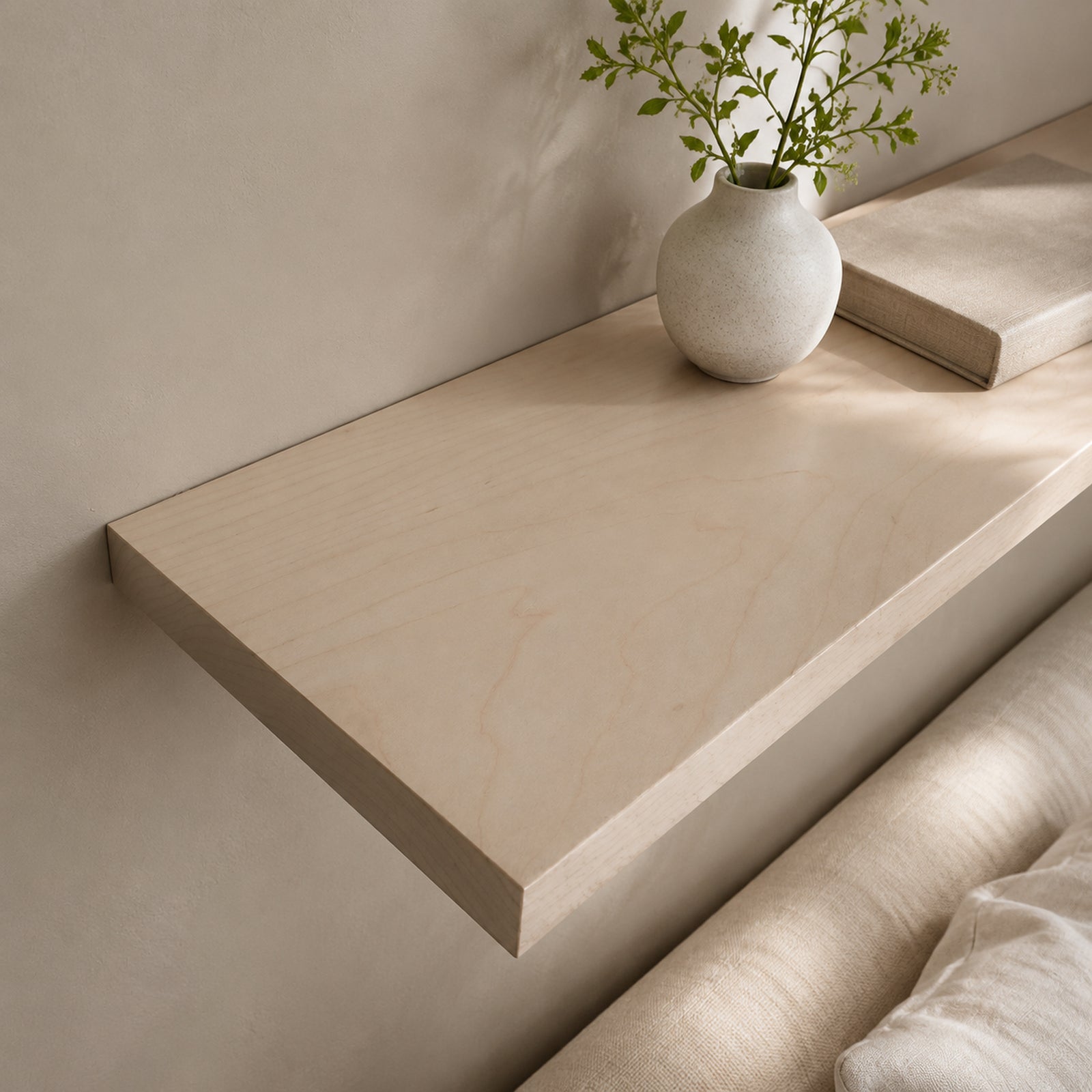 A light beige floating shelf mounted on a neutral wall, styled with a small textured white vase holding green stems and a closed book, with part of a beige upholstered chair visible in the foreground.