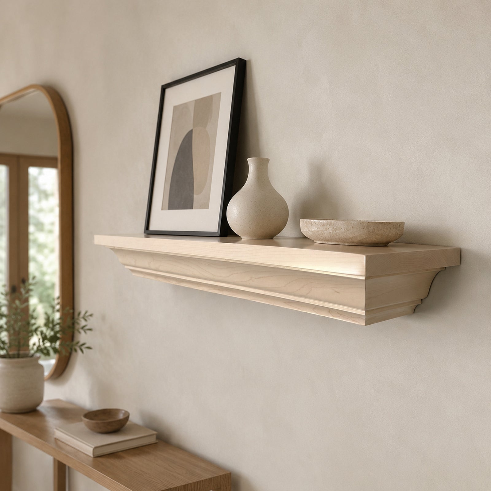 A light natural wood floating shelf mounted on a textured beige wall, styled with a black-framed abstract print, a beige ceramic vase, and a matching shallow bowl. A wooden console table and a mirror are partially visible in the softly lit room.