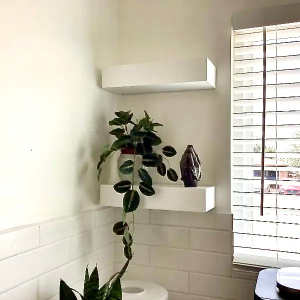 White floating wall shelves mounted on a bathroom wall, with a trailing green houseplant and a dark vase displayed on the shelves beside a window with blinds.