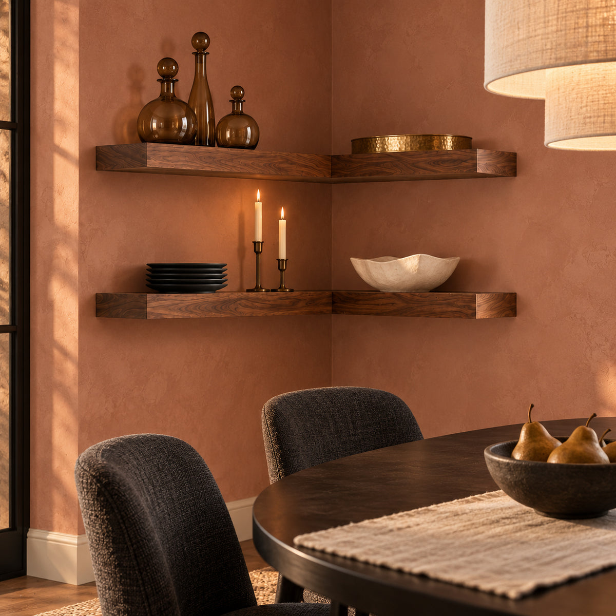 A styled dining room corner with two dark wood floating corner shelves mounted on a terracotta-colored wall. The shelves are decorated with amber glass bottles, plates, candles, and a white bowl, with a dark wood dining table, textured chairs, and a hanging lamp visible in the foreground.