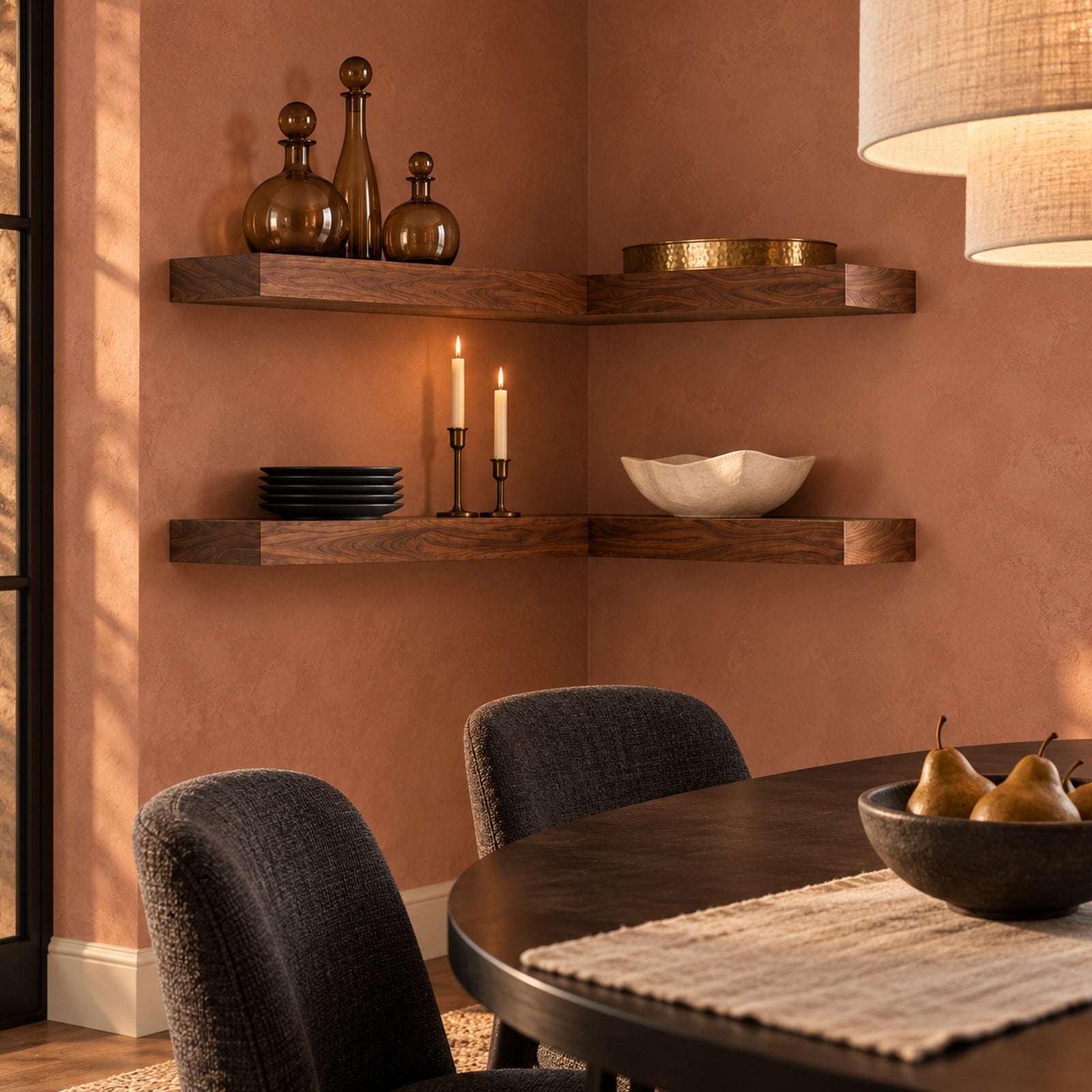 A styled dining room corner with two dark wood floating corner shelves mounted on a terracotta-colored wall. The shelves are decorated with amber glass bottles, plates, candles, and a white bowl, with a dark wood dining table, textured chairs, and a hanging lamp visible in the foreground.