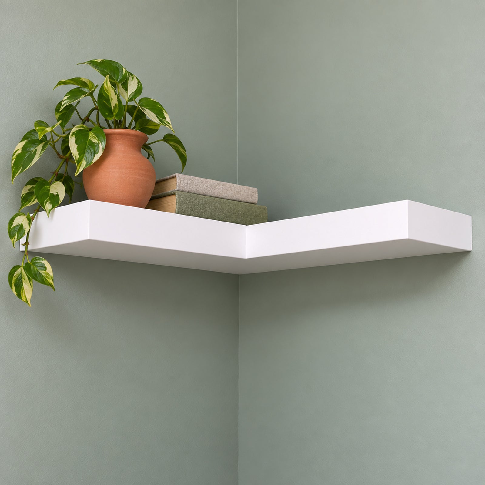 A modern white floating corner shelf mounted on a sage green wall, styled with a terracotta planter and books.
