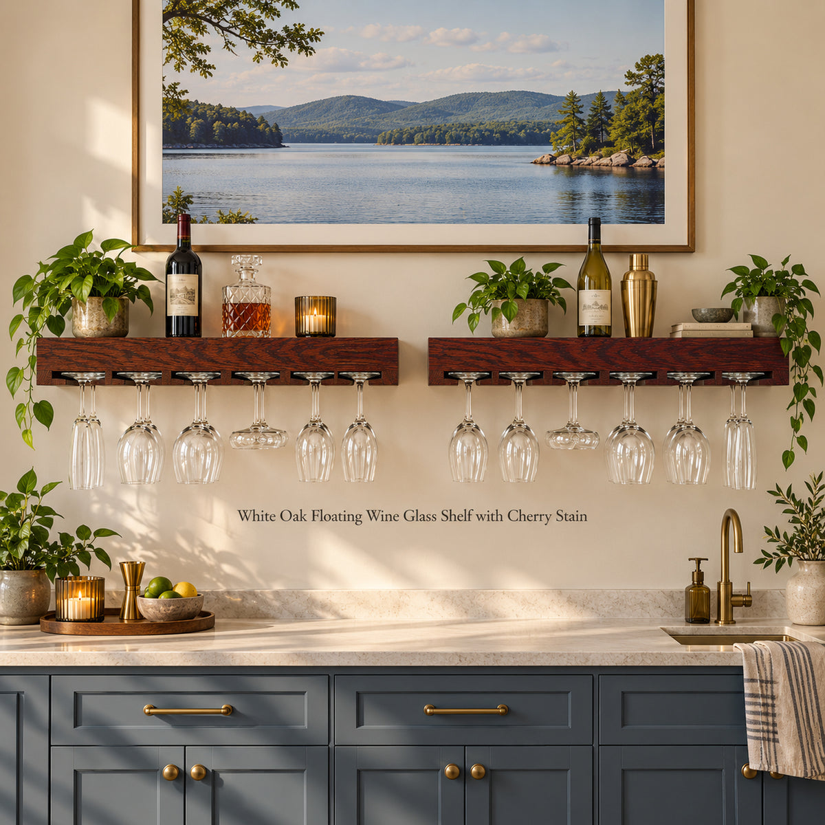 Two dark wood floating wine glass holders mounted on a beige wall above a kitchen counter, with wine glasses and other stemware hanging from each shelf. The scene includes potted plants, bottles, candles, a framed lake landscape print, blue-gray cabinets, and a gold faucet.