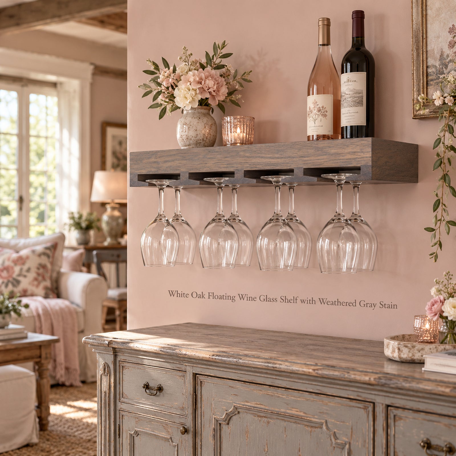 A floating wine glass holder shelf mounted on a pink wall in a styled living room. The shelf is made of white oak with a weathered gray stain and holds eight hanging wine glasses.