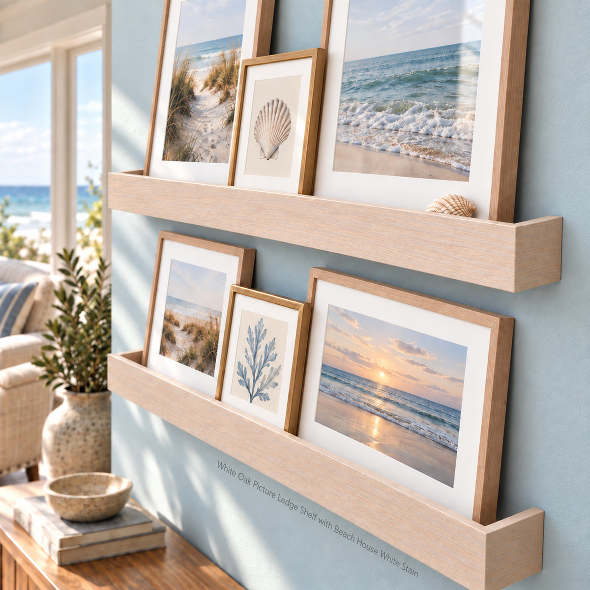 Two light wood floating shelves mounted on a pale blue wall, styled with framed beach and dune photographs. A small shell rests on the upper shelf, and a vase with greenery and books are visible on a wooden console table below. Text reads 'White Oak Picture Ledge Shelf with Beach House White Stain'.