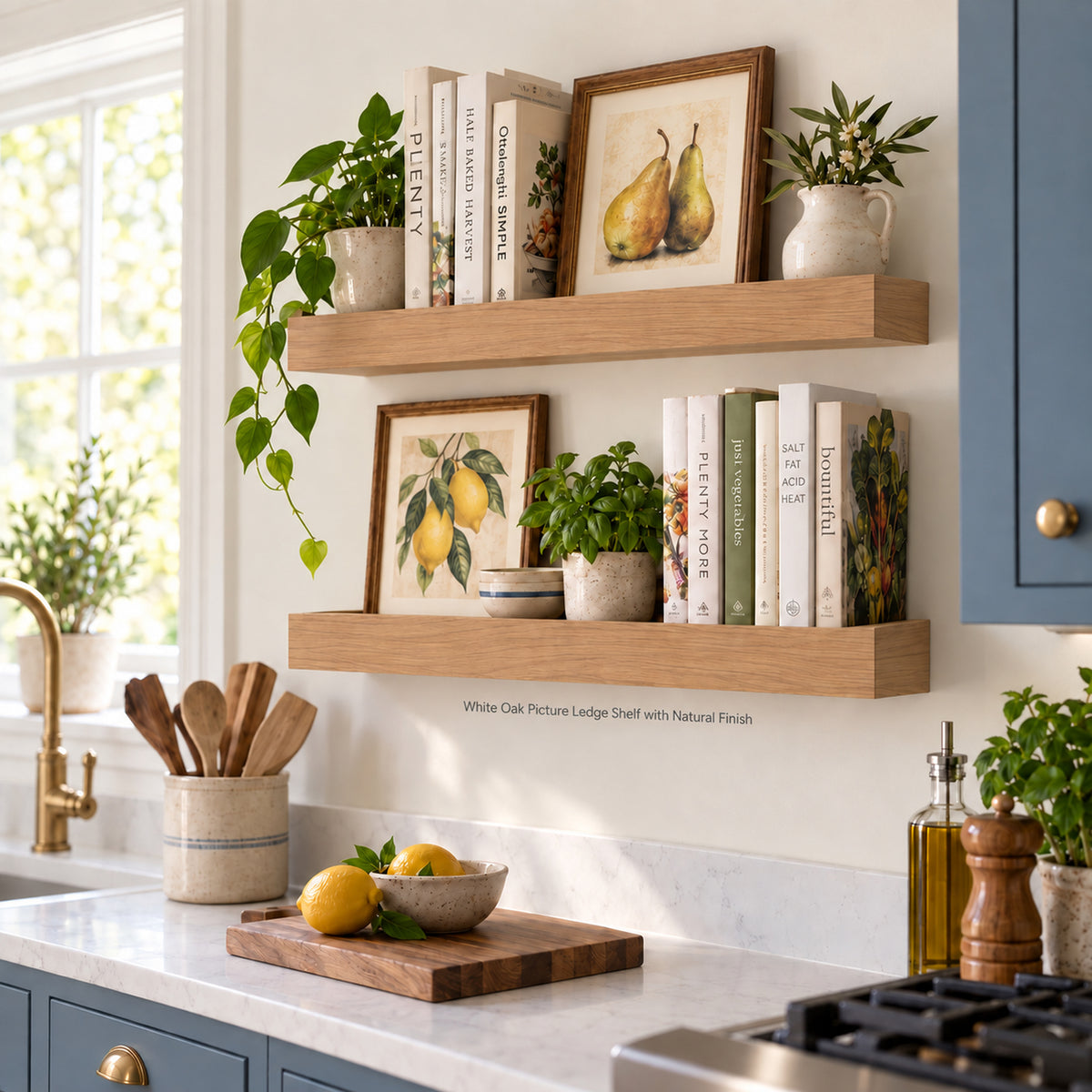 Two white Oak picture ledge shelves mounted on a kitchen wall, styled with cookbooks, framed pear and lemon prints, small potted plants, and ceramic dishes. The kitchen features blue cabinets, a white countertop, and a brass faucet.